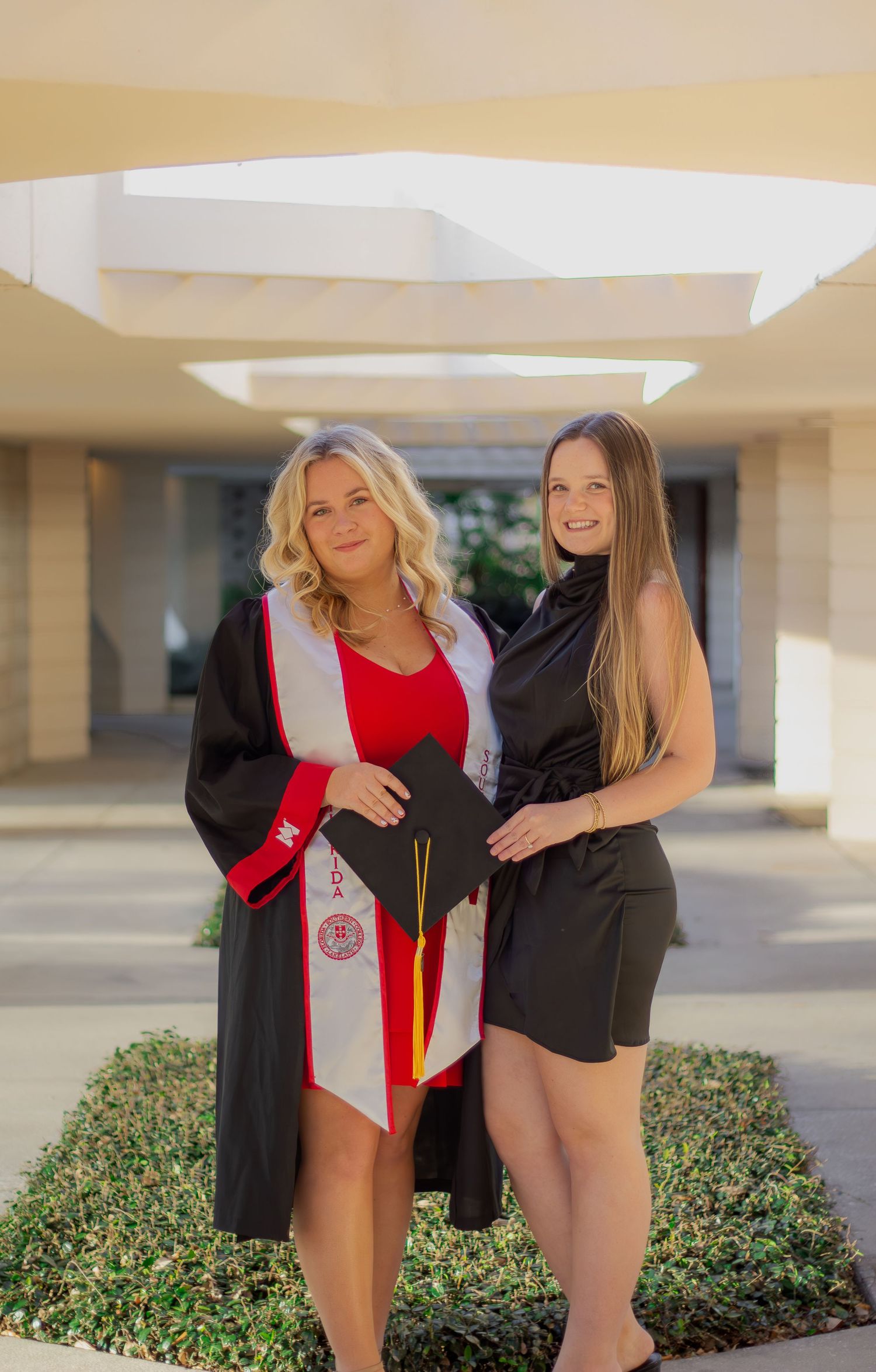Two women pose together outdoors, one wearing a graduation gown with red stole and the other in a black dress.