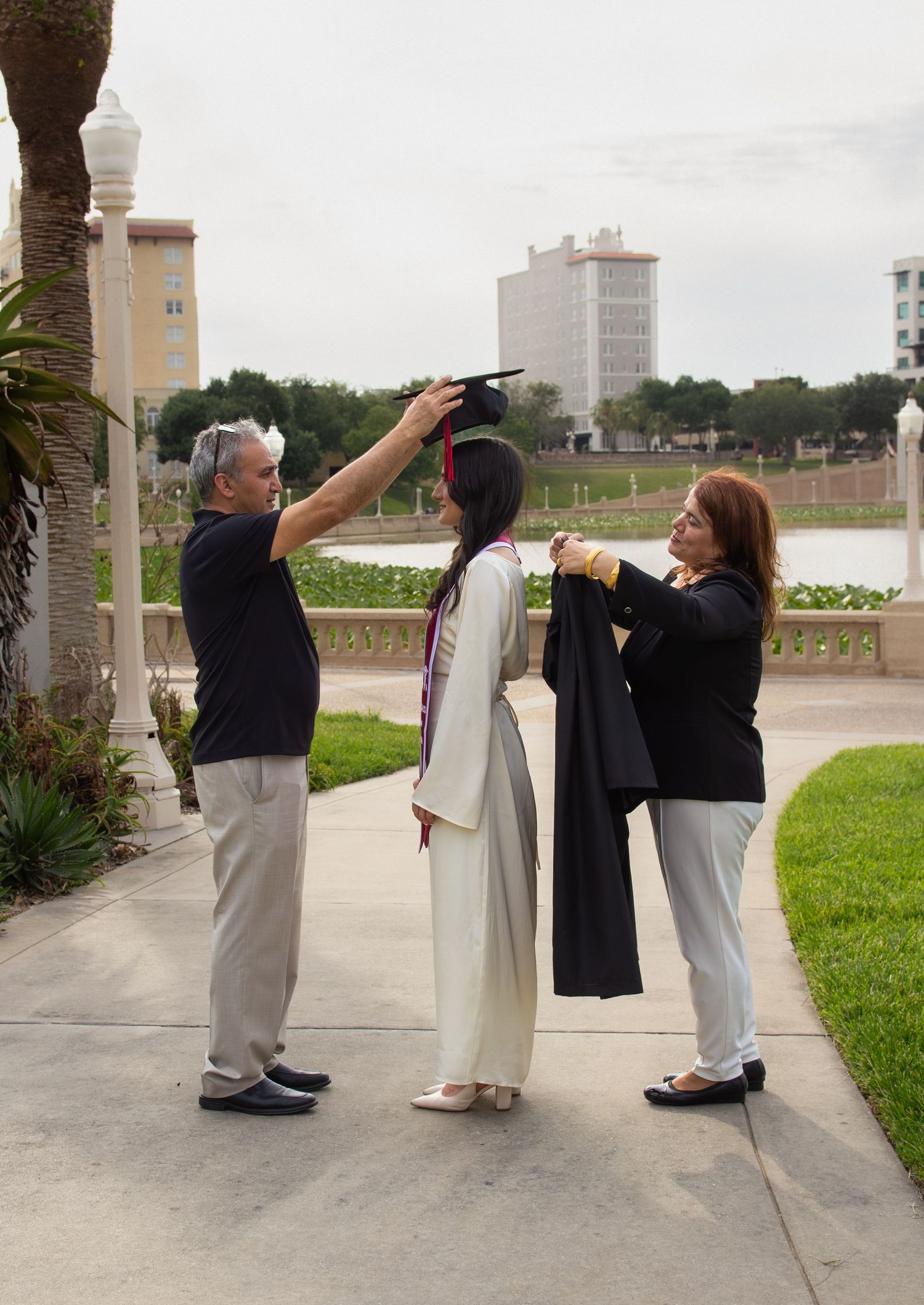 Parents helping their graduate daughter put on cap and gown outdoors on a college campus.