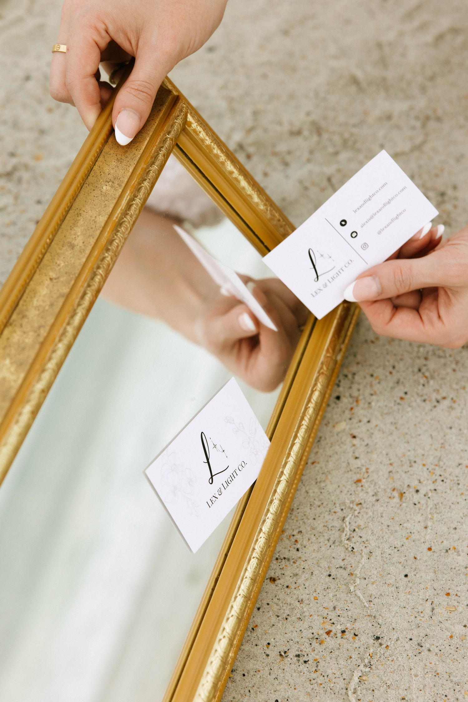 Hands holding a white business card near a gold ornate mirror reflecting the card's design on concrete.