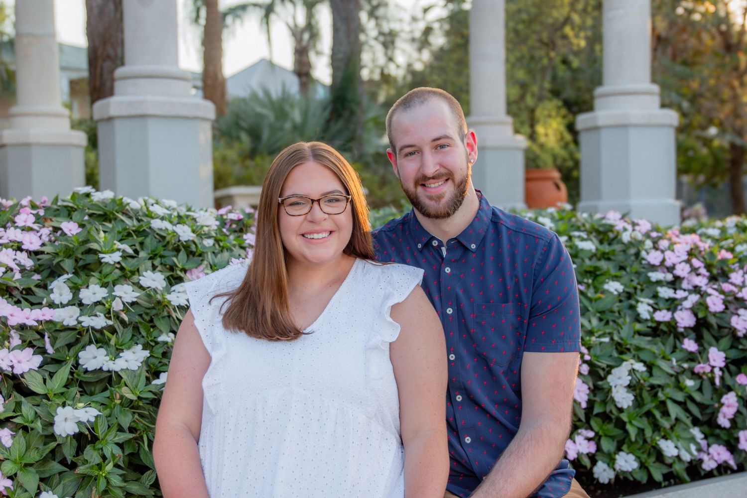 Happy couple poses together by white garden columns surrounded by pink flowers.