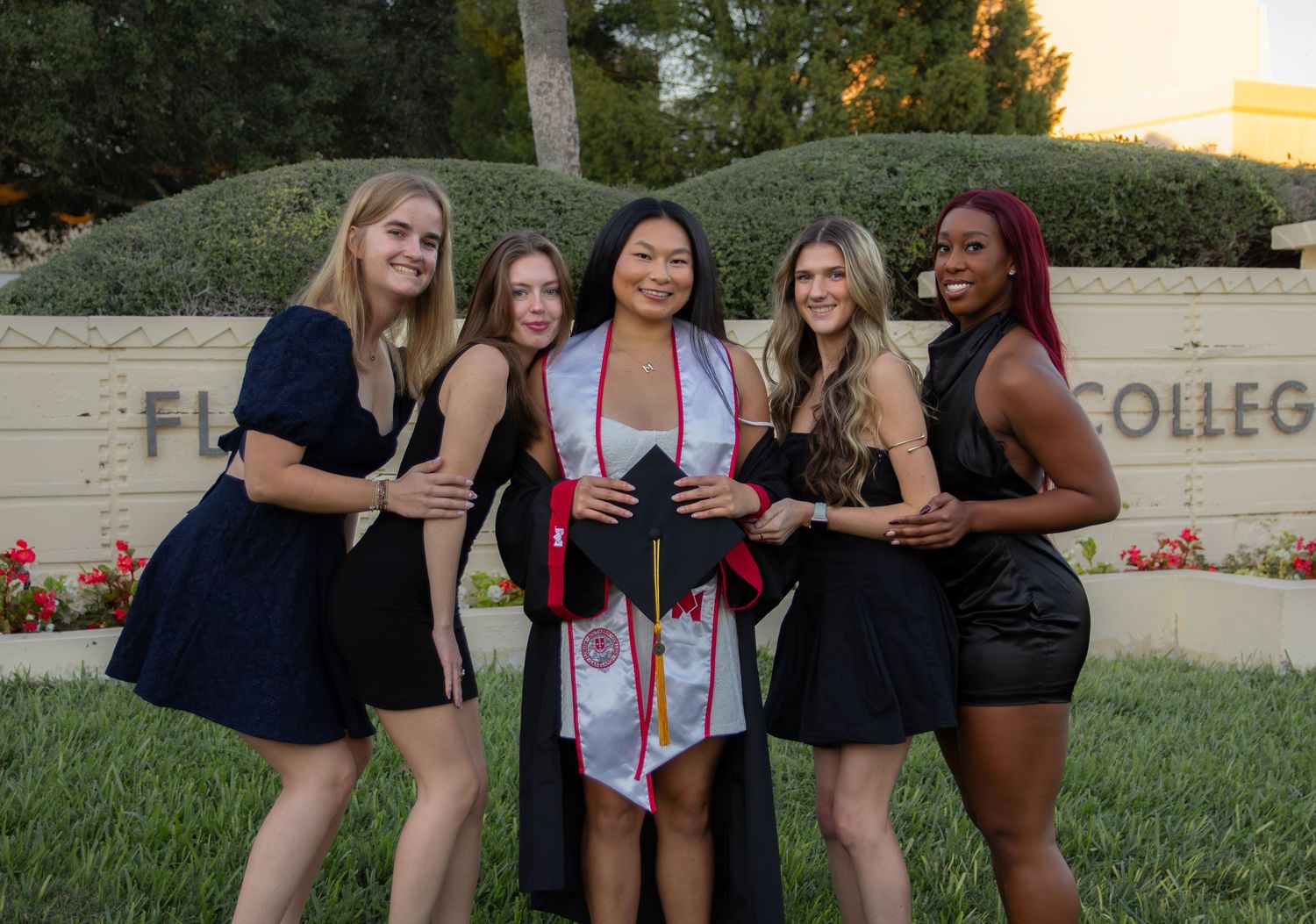 A group of friends in formal dresses pose together on a grassy lawn in front of a college sign at sunset.