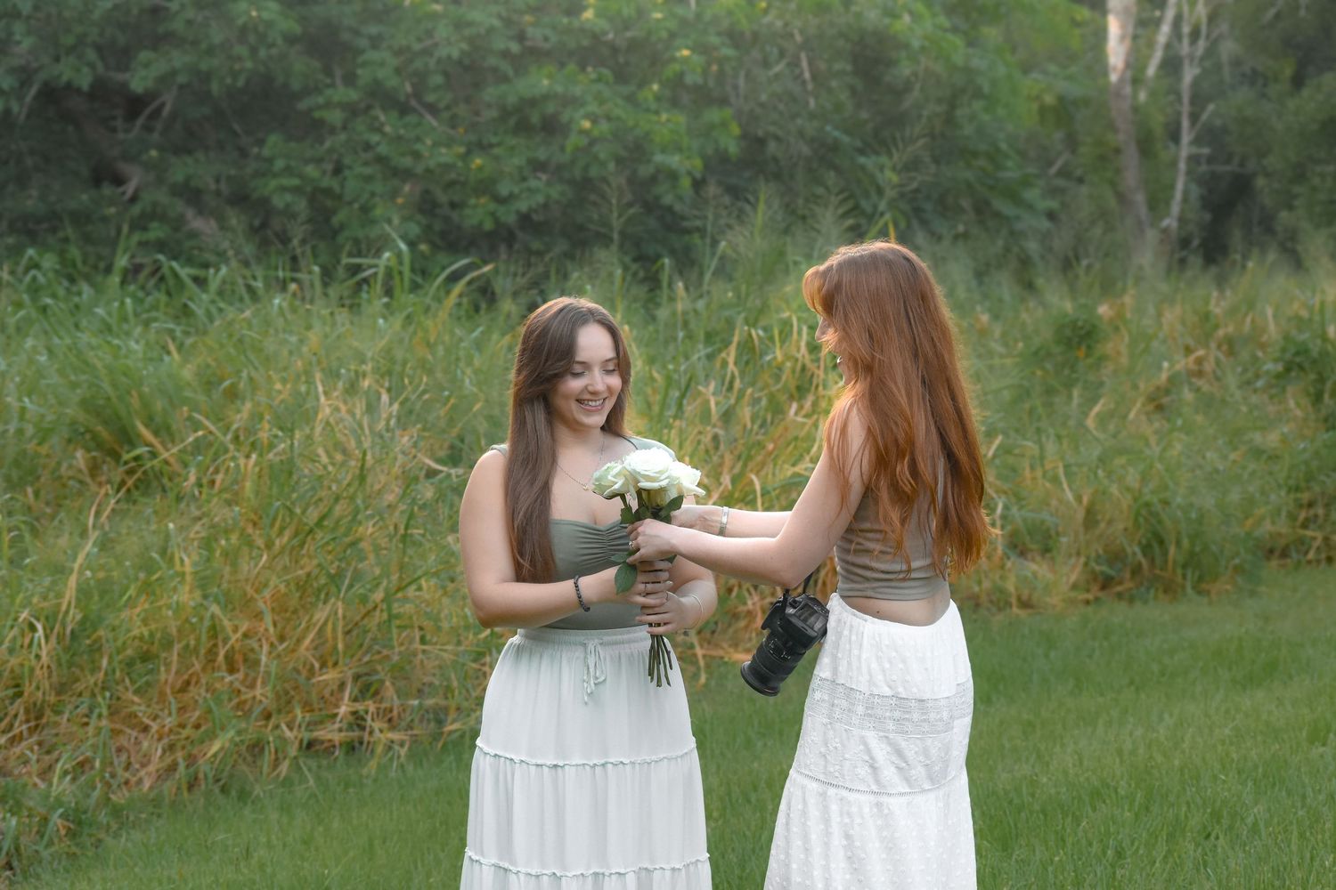 Two people in white dresses sharing flowers while standing in a lush green outdoor setting.