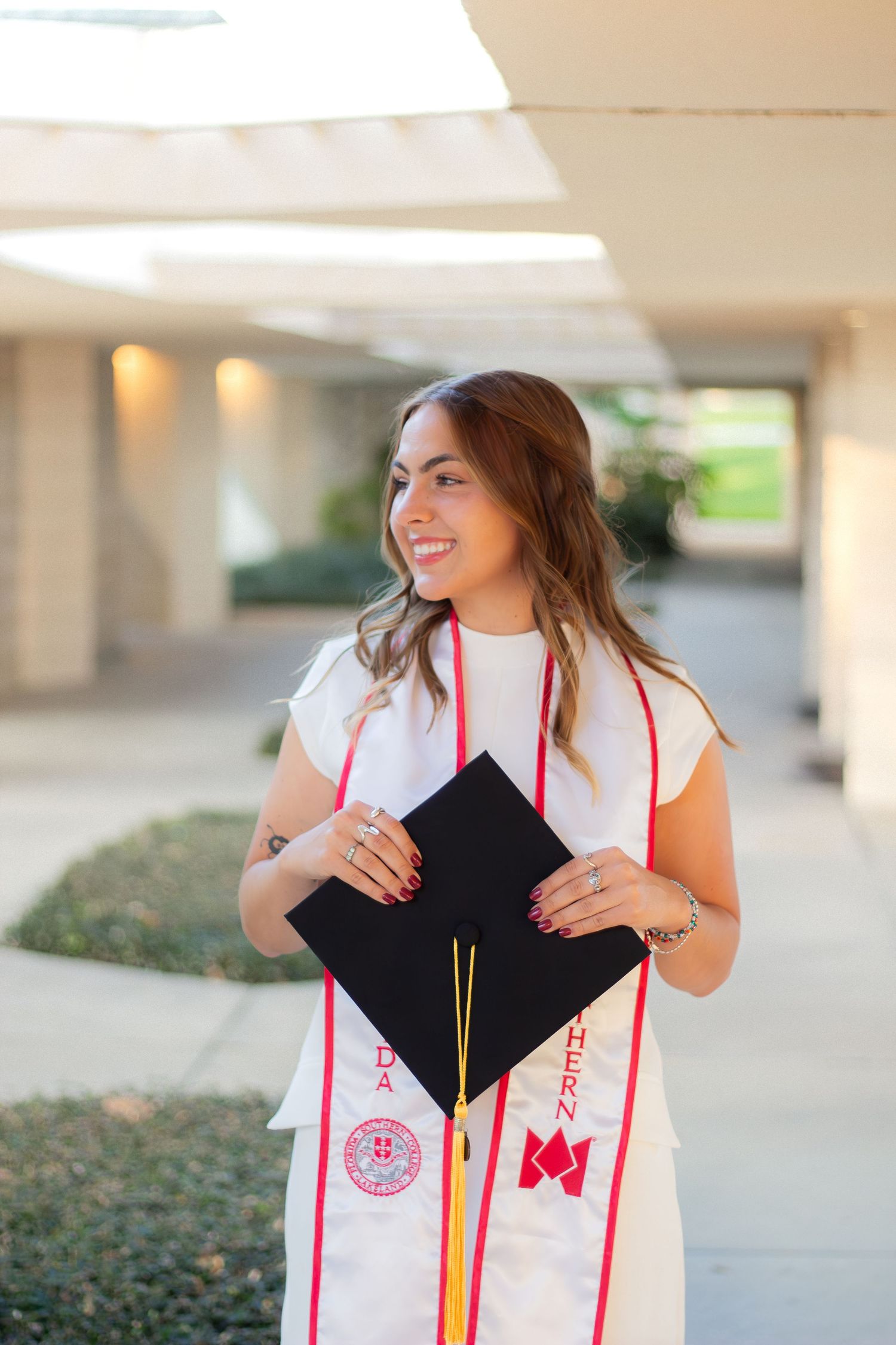 Graduate wearing white stole and holding black mortarboard cap in outdoor graduation photoshoot setting.