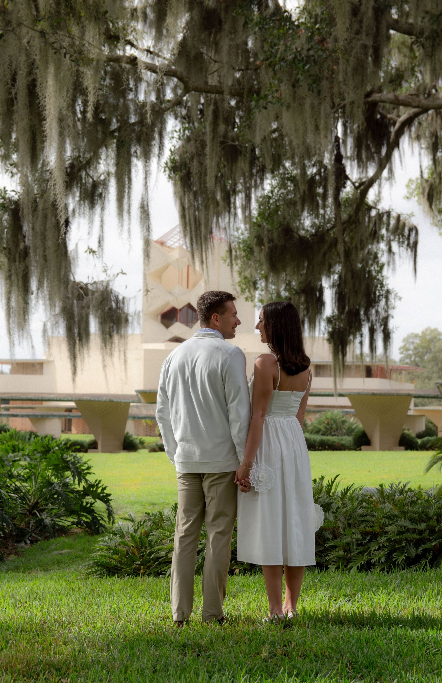 A couple stands together under Spanish moss draped trees in a romantic outdoor setting.