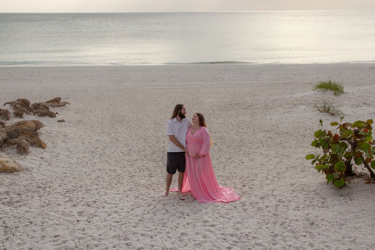 Couple stands on sandy beach at sunset, woman in flowing pink maternity gown, ocean waves in background.