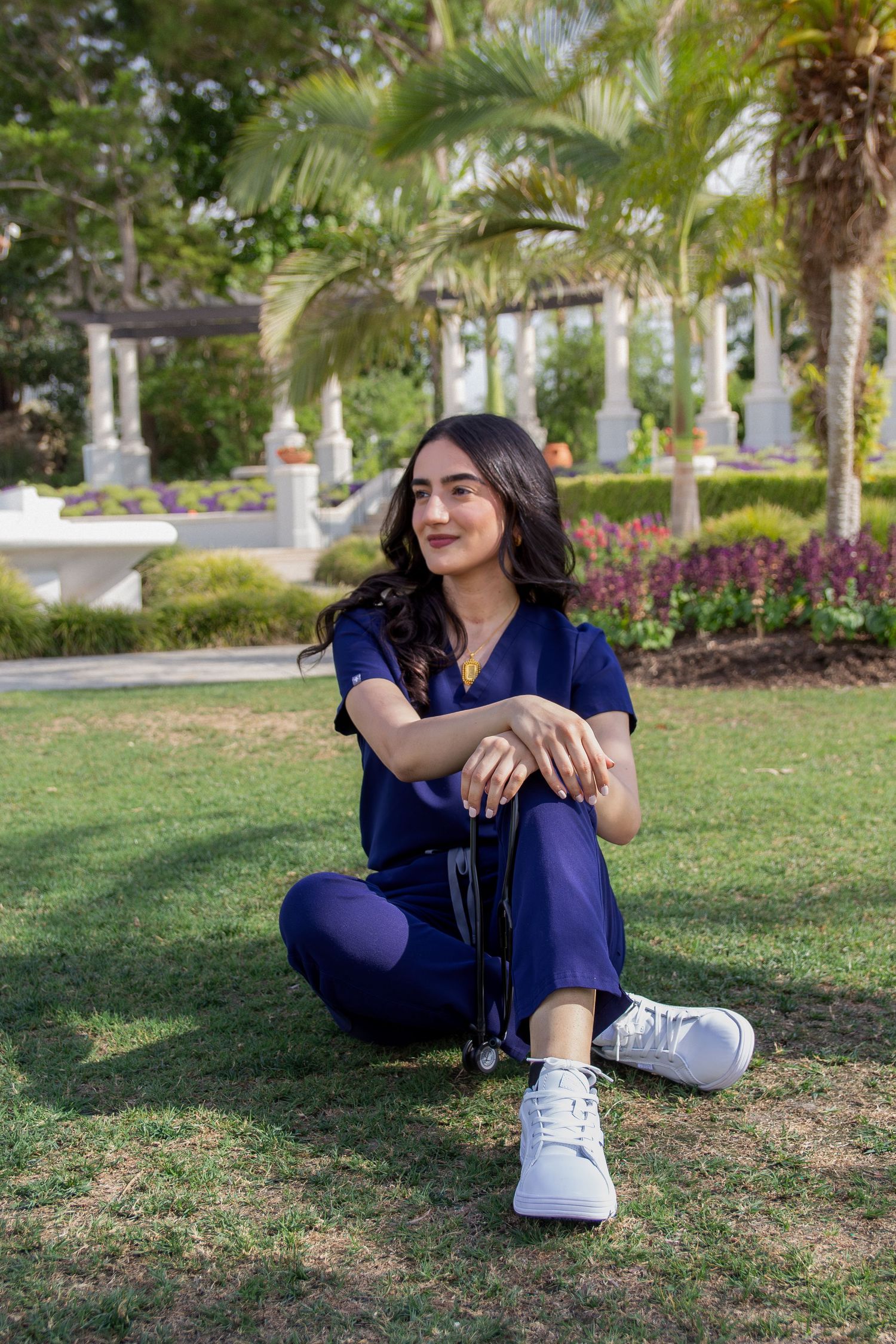 Young woman in navy blue scrubs sitting on grass in a lush garden with palm trees and white columns.