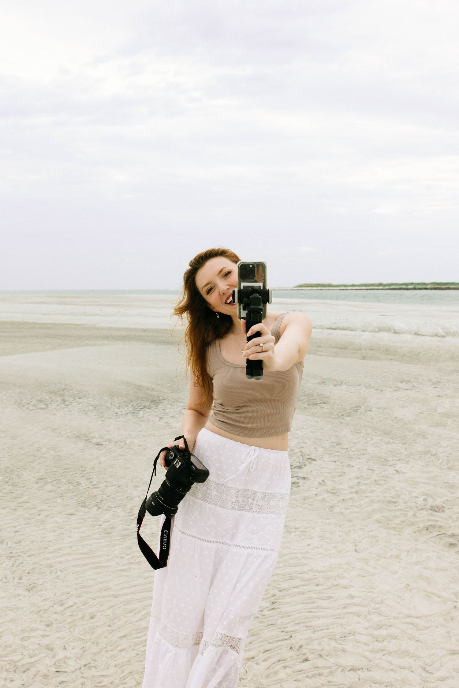 Smiling woman holding a camera on a beach, wearing a tan top and white skirt on a cloudy day.
