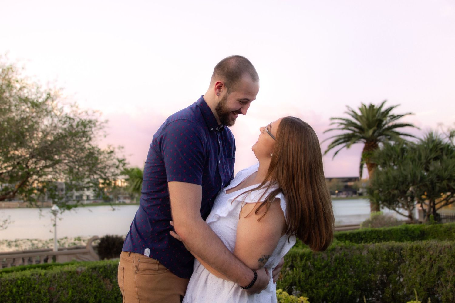 A romantic couple embraces during a sunset photoshoot in a tropical outdoor setting with palm trees.