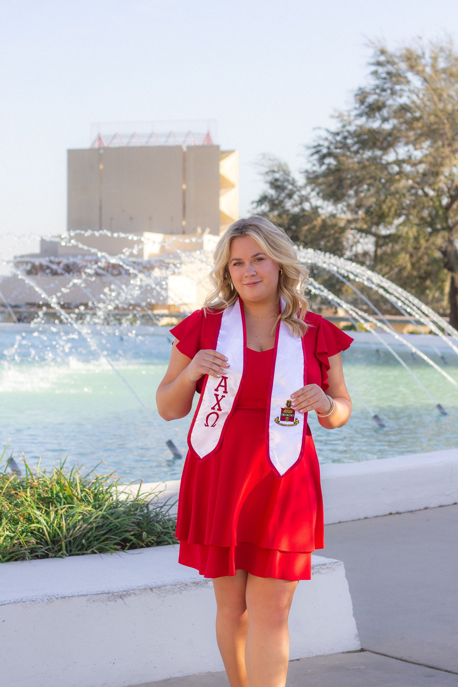 Graduate in a red dress and white sash poses in front of a fountain on a sunny day at university.