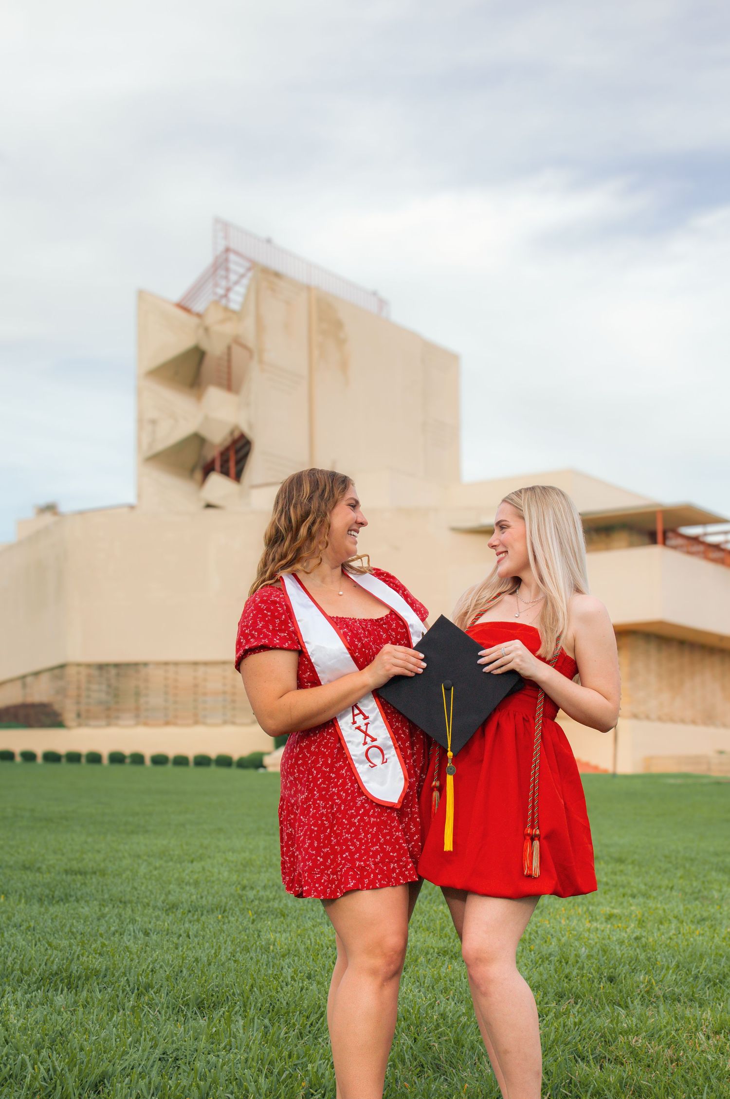 Two friends in red outfits pose playfully together on a grassy field with buildings in the background.