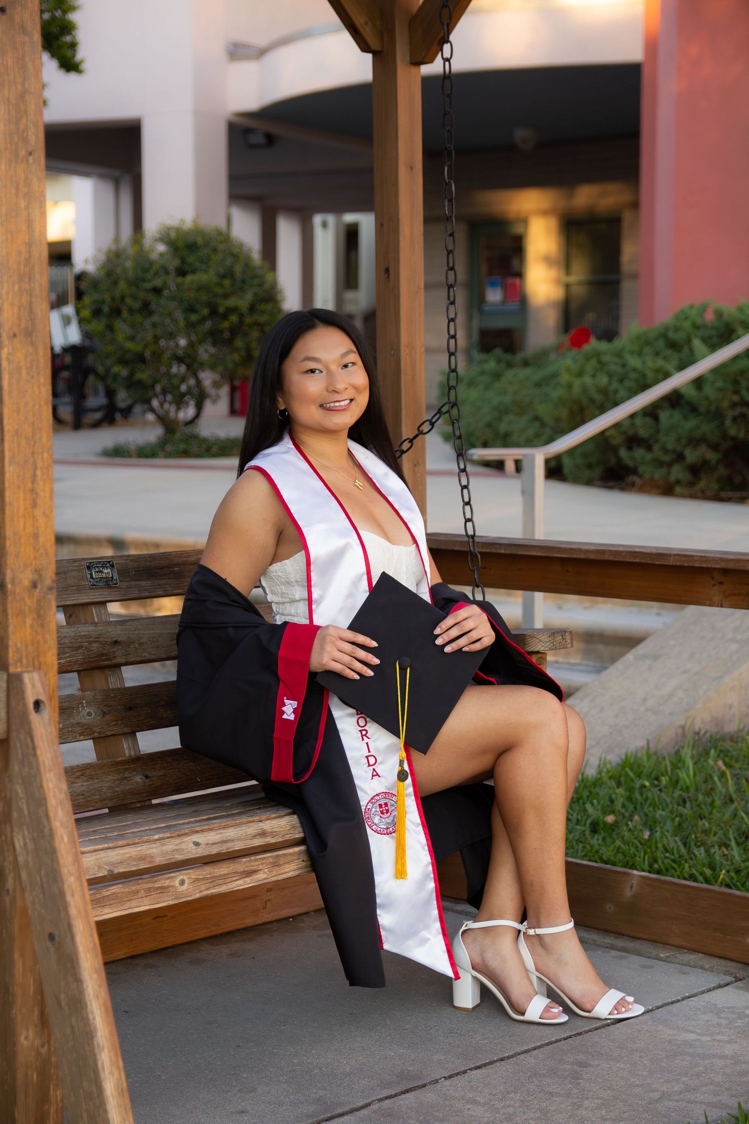 A person sits on a wooden swing bench wearing a black dress and white sandals in an outdoor courtyard.