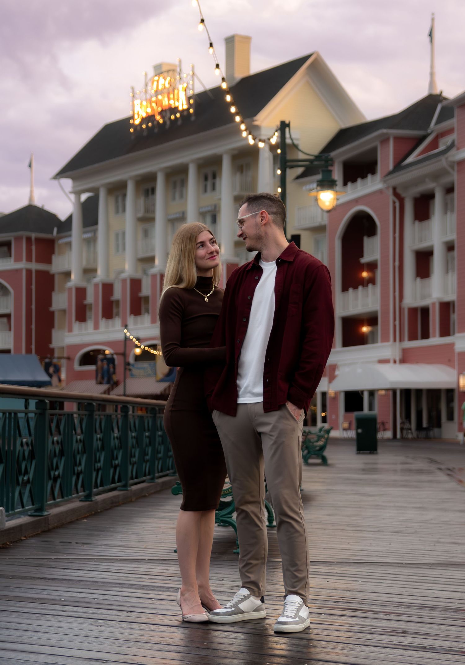 A couple shares a romantic moment on a Victorian-style boardwalk at dusk with string lights overhead.