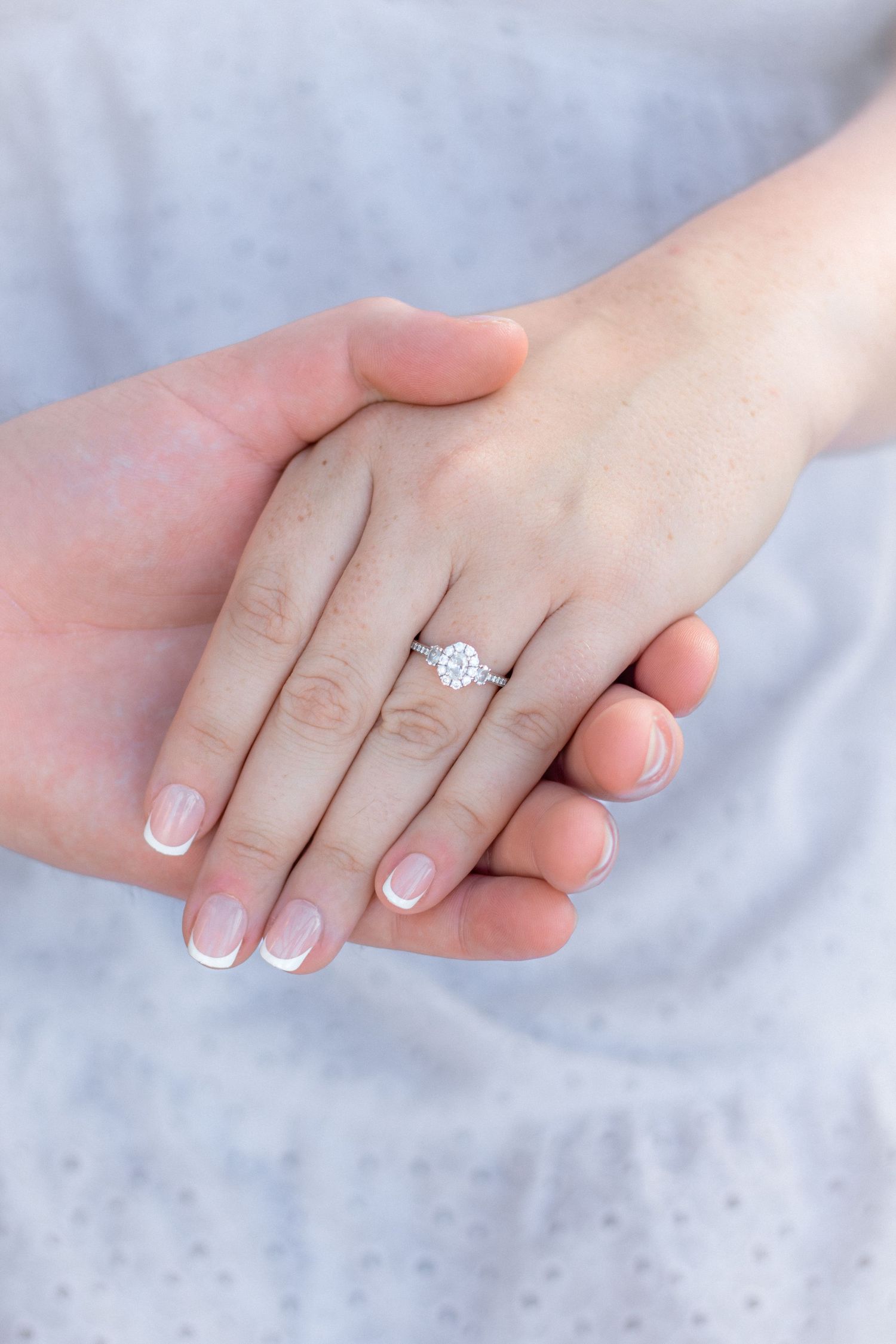 Close-up engagement ring shots against white fabric showing delicate diamond solitaire and french manicured hands.