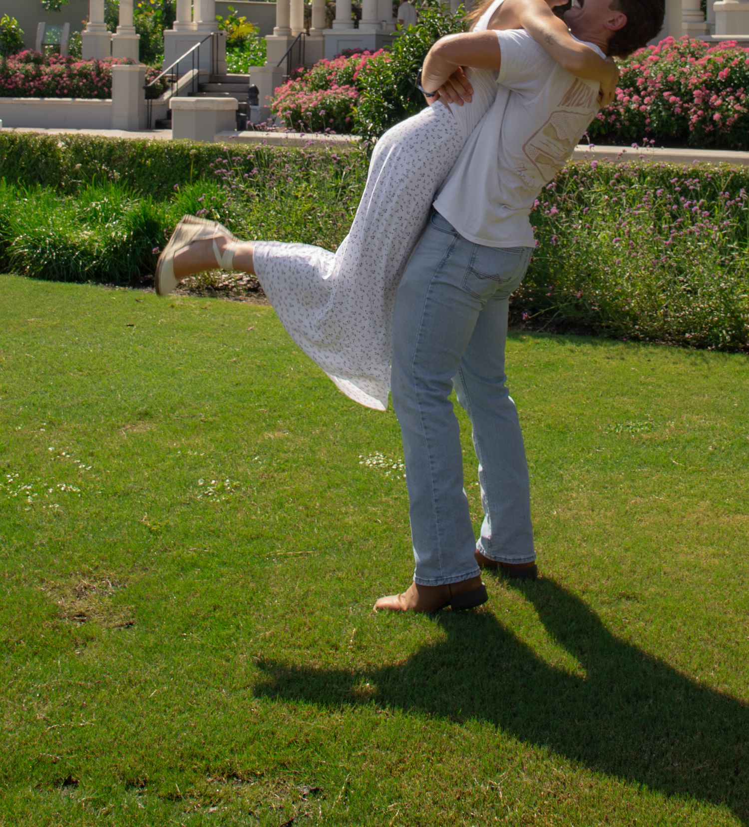 Couple embracing and lifting each other joyfully on a sunny green lawn outside a elegant building.