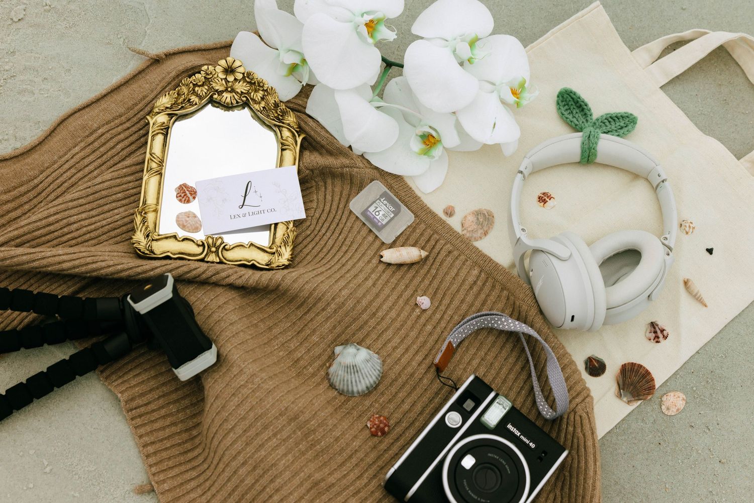 Flat lay of white headphones, vintage camera, gold mirror, orchids, and brown knit sweater on a neutral background.