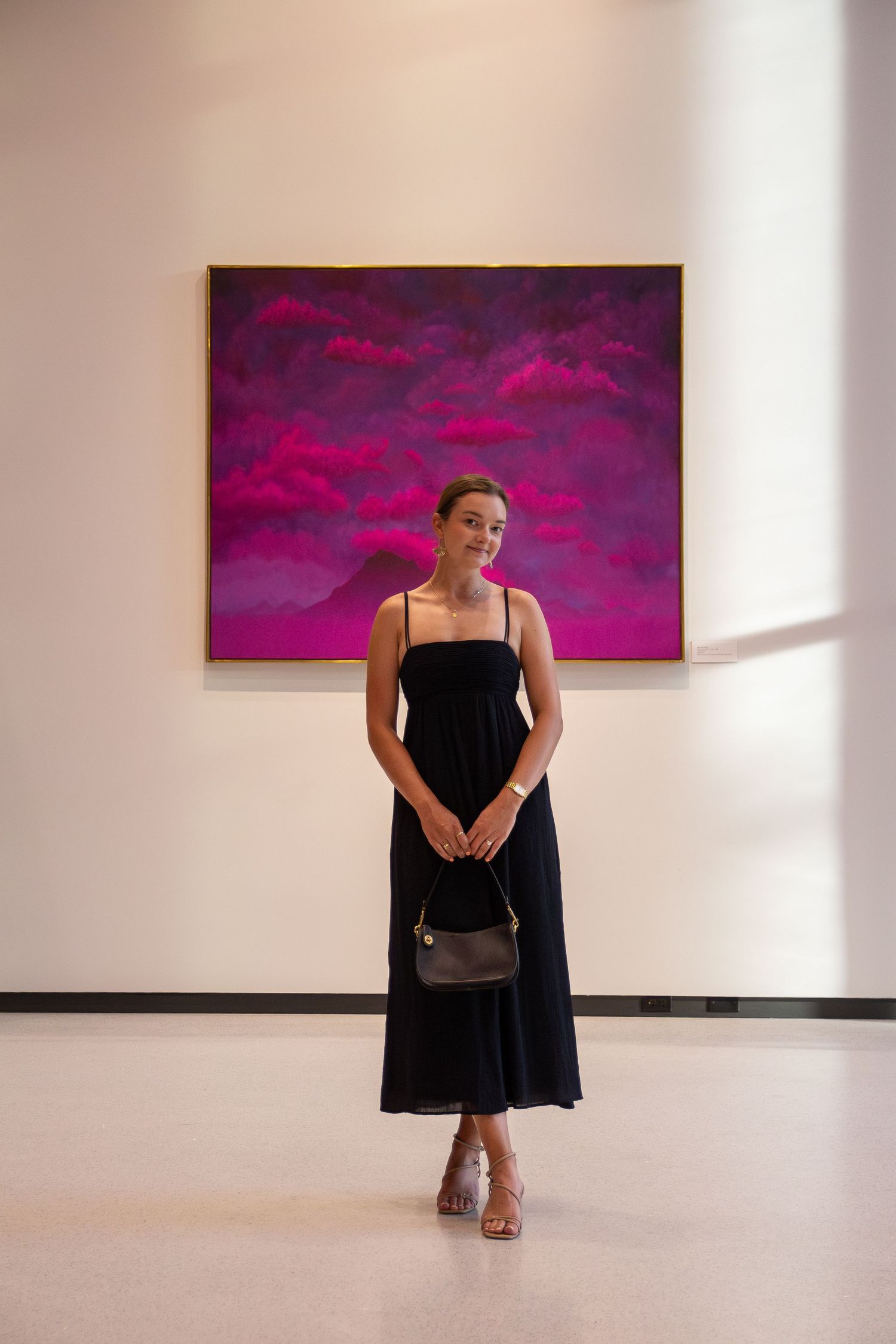 A person wearing a black dress stands near a magenta artwork in a minimalist white gallery.