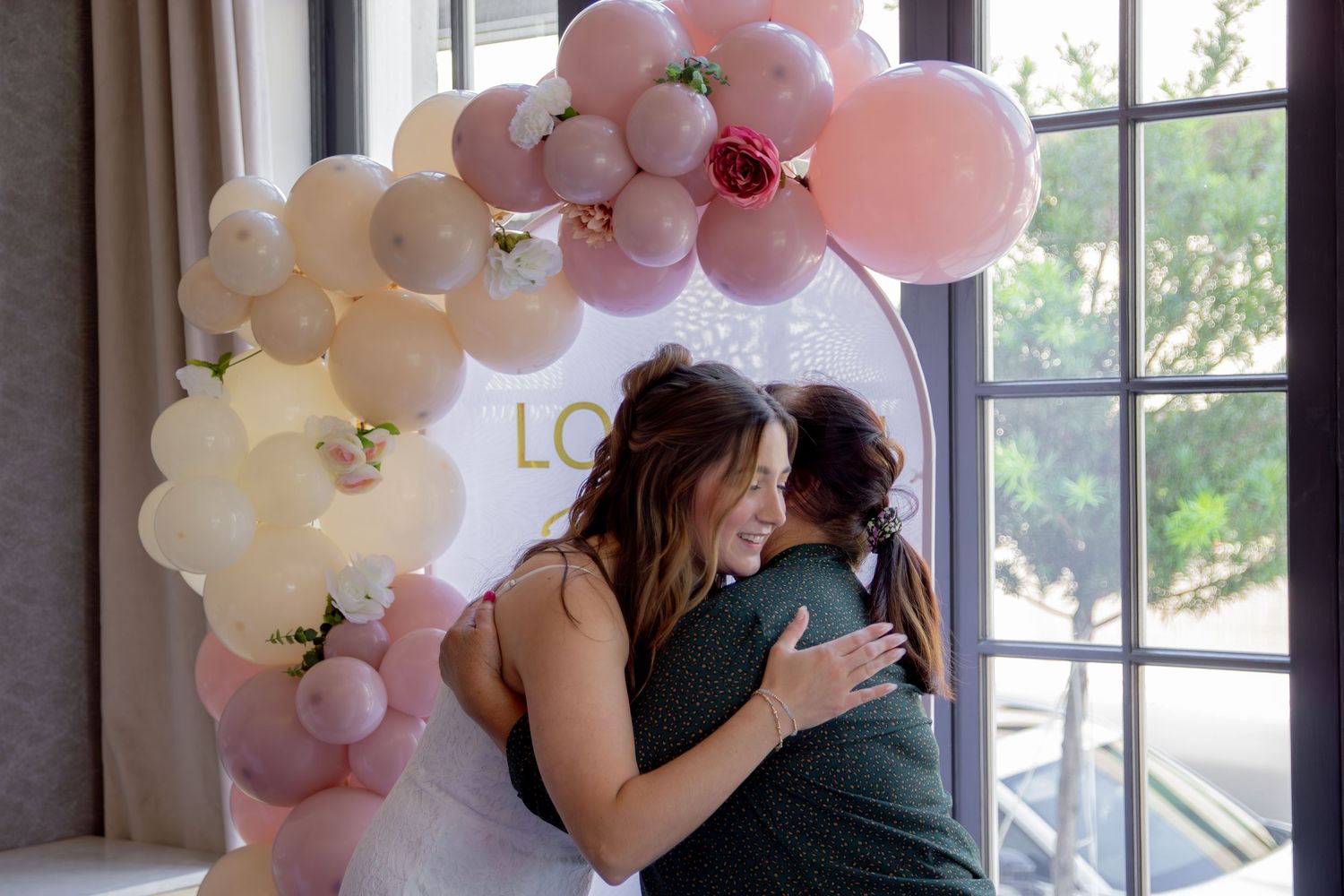 A couple embraces during a romantic moment surrounded by pink and white balloon decorations near a window.