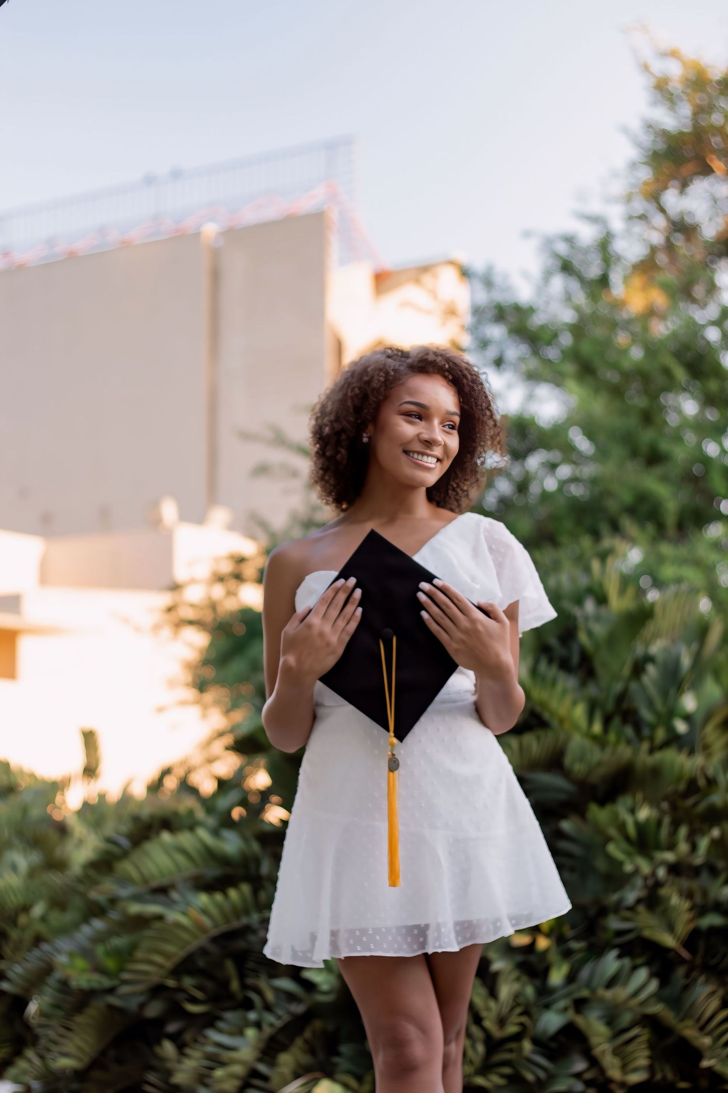 A graduate in a white dress and graduation cap poses outdoors against greenery during a celebratory photoshoot.