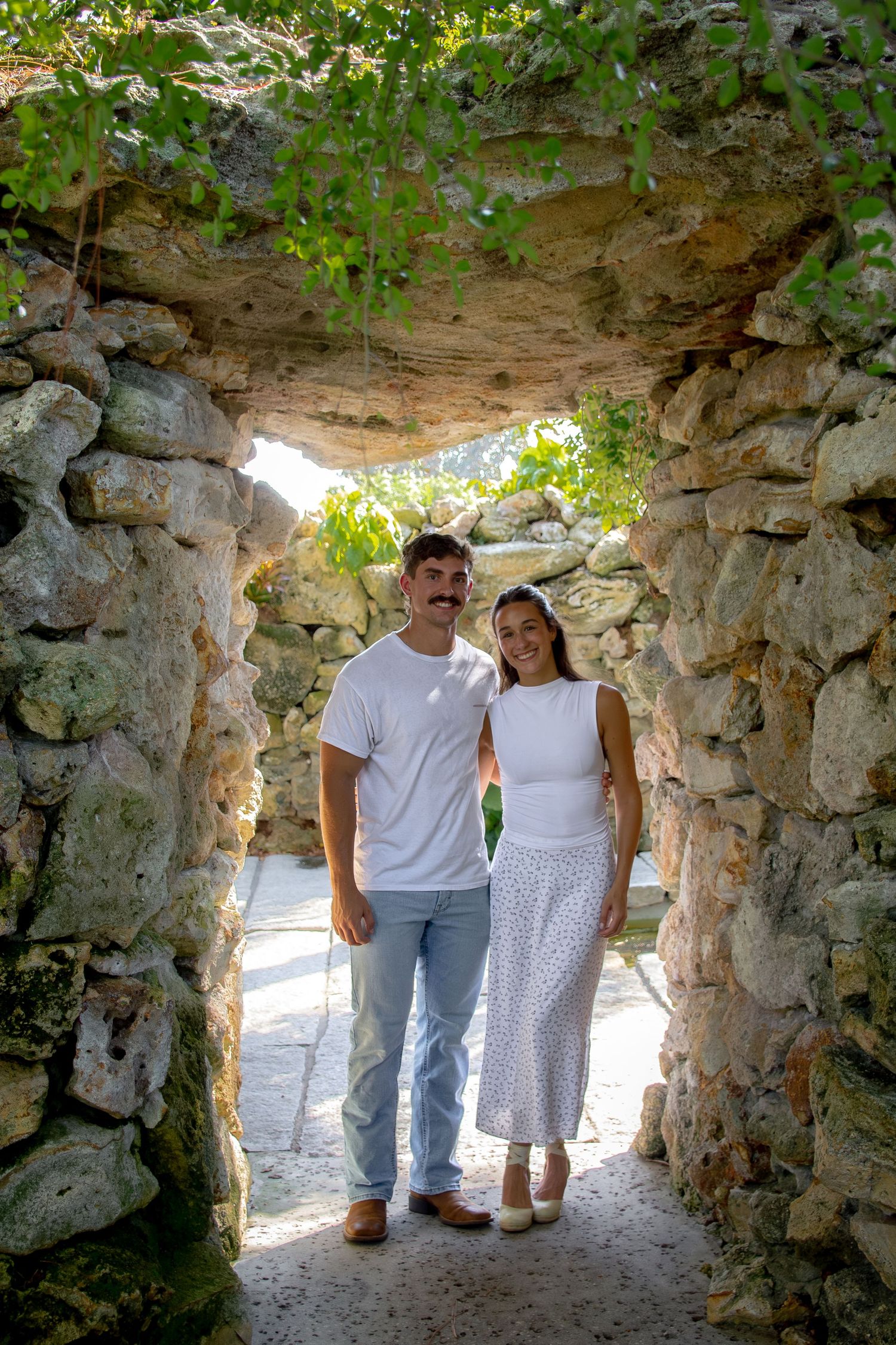 Couple in white outfits posing under a rustic stone archway surrounded by lush greenery on a sunny day.
