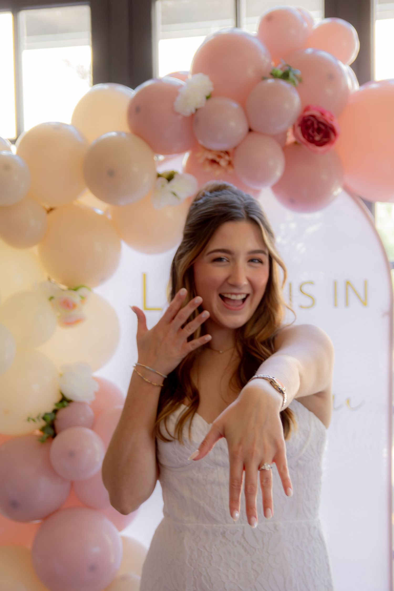 A bride in a white lace dress shows off her ring in front of a pink and white balloon arch backdrop.
