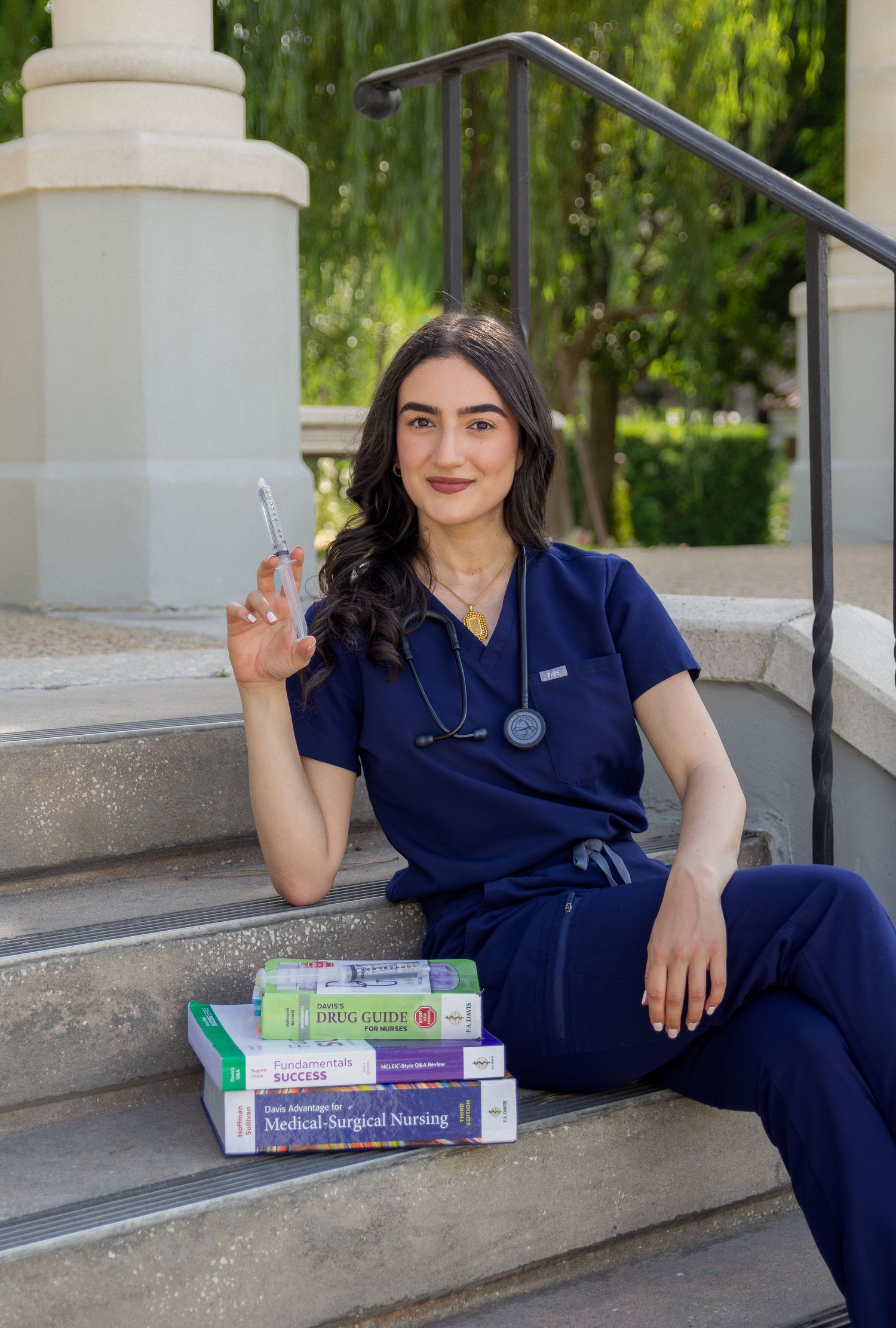 Nursing student in navy scrubs sitting on steps with medical textbooks and stethoscope, holding scissors outdoors.