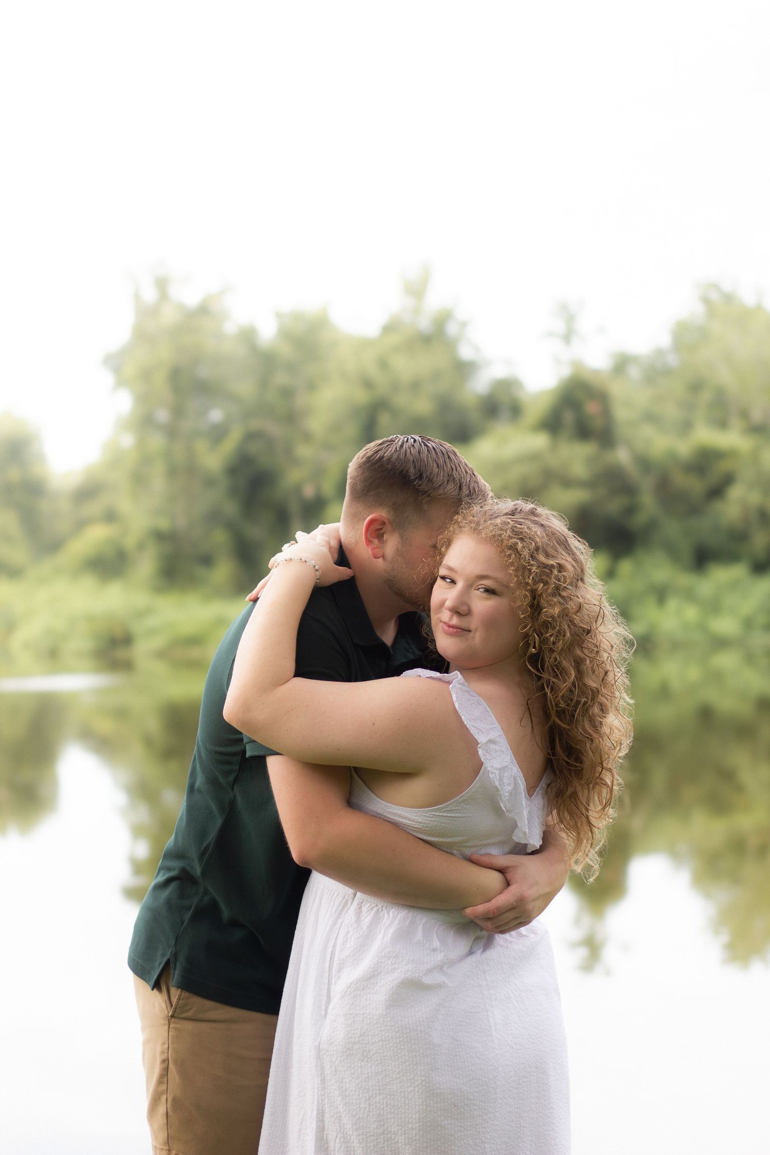 A tender moment captured between a couple embracing by a tranquil lake surrounded by greenery.