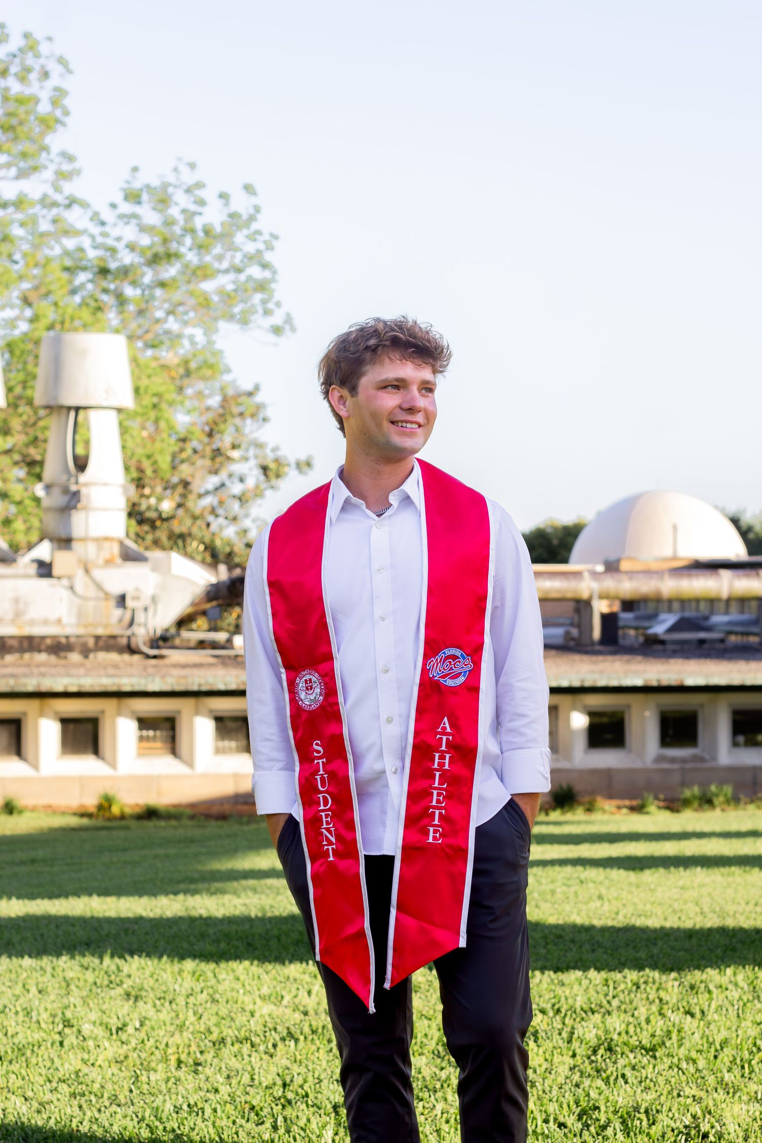 Graduate wearing a red and white stole stands on grass with buildings in background for outdoor portraits.