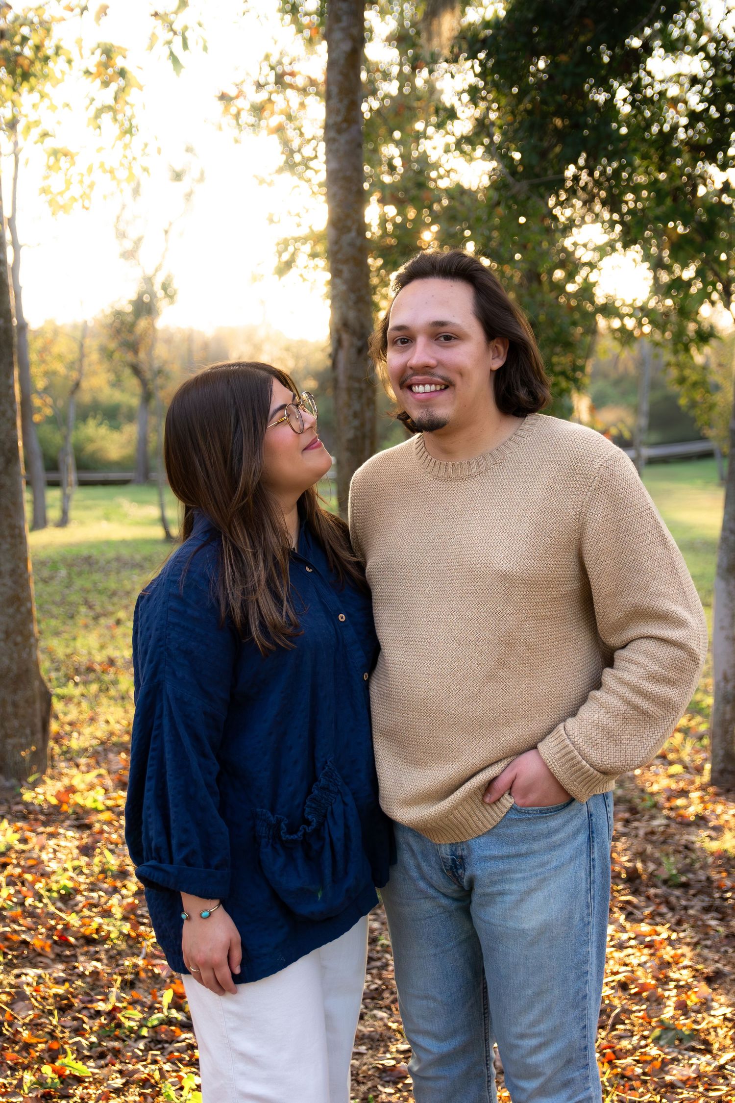 A couple shares a tender moment in a park during autumn with fallen leaves on the ground and golden sunlight.