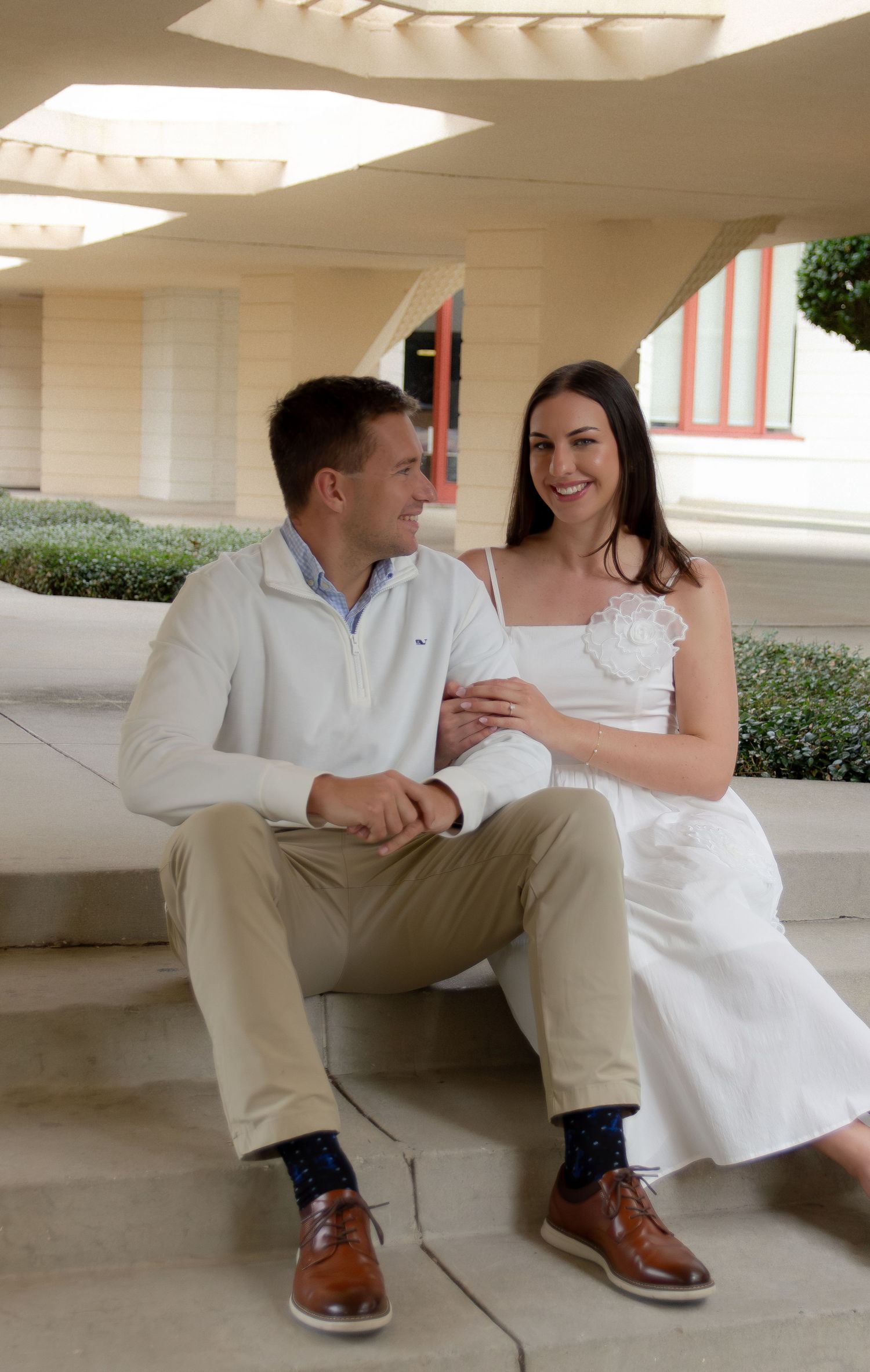 Couple sitting together in a modern architectural setting wearing white and khaki casual attire.