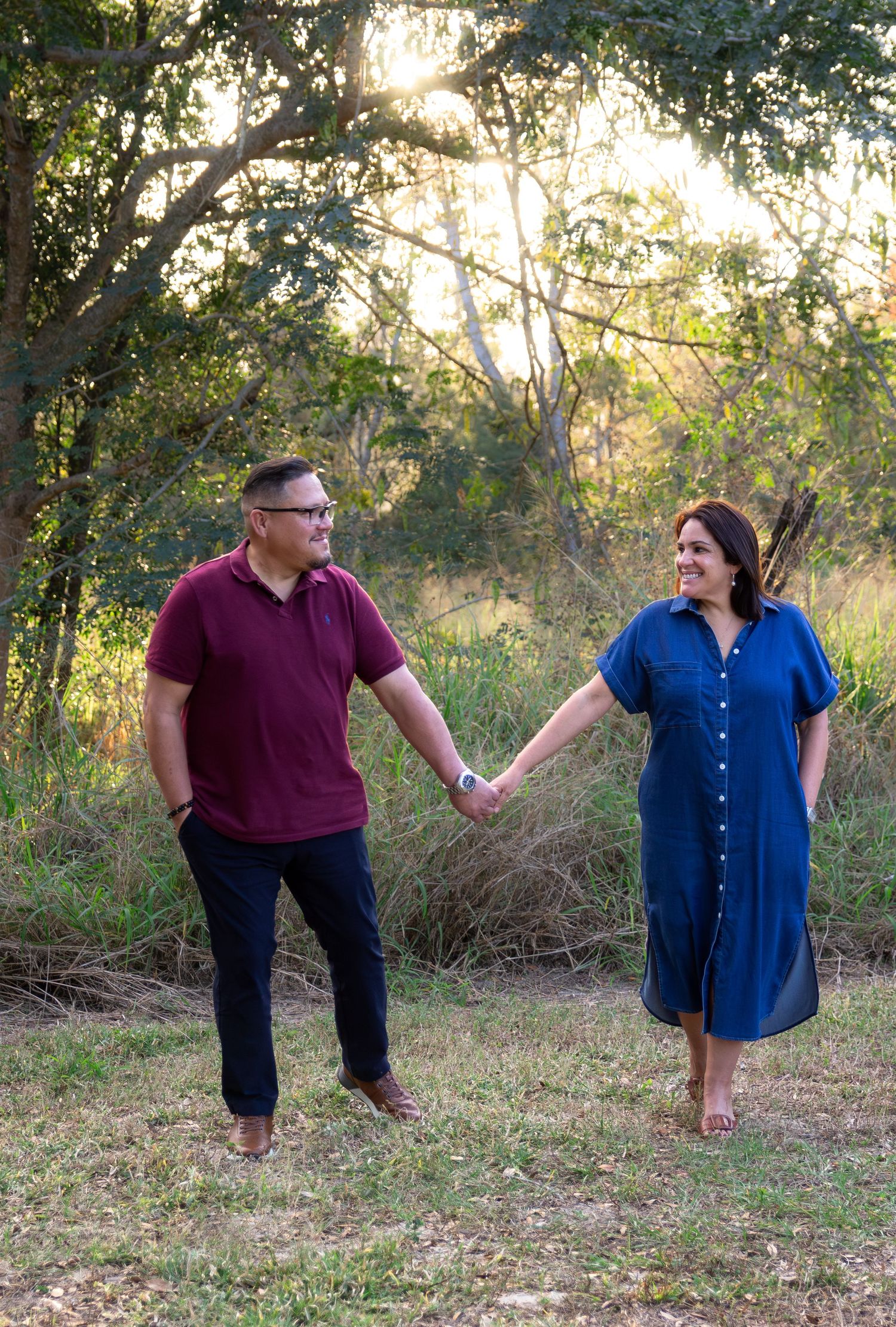 A couple in burgundy and blue outfits playfully interacting in a grassy outdoor setting at sunset.