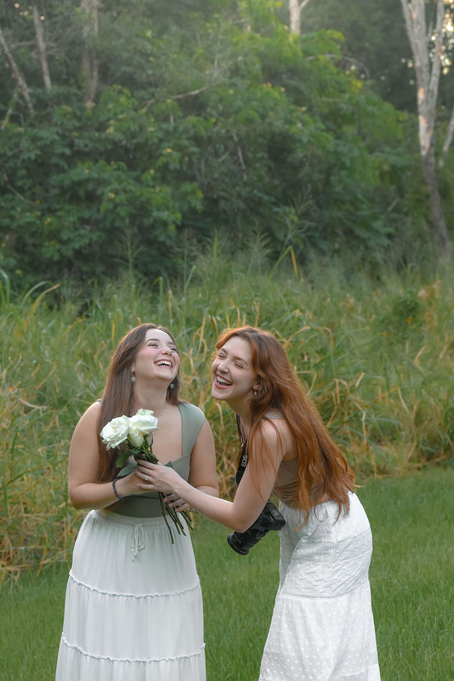 Two people in white dresses laugh together while holding flowers in a grassy outdoor field.