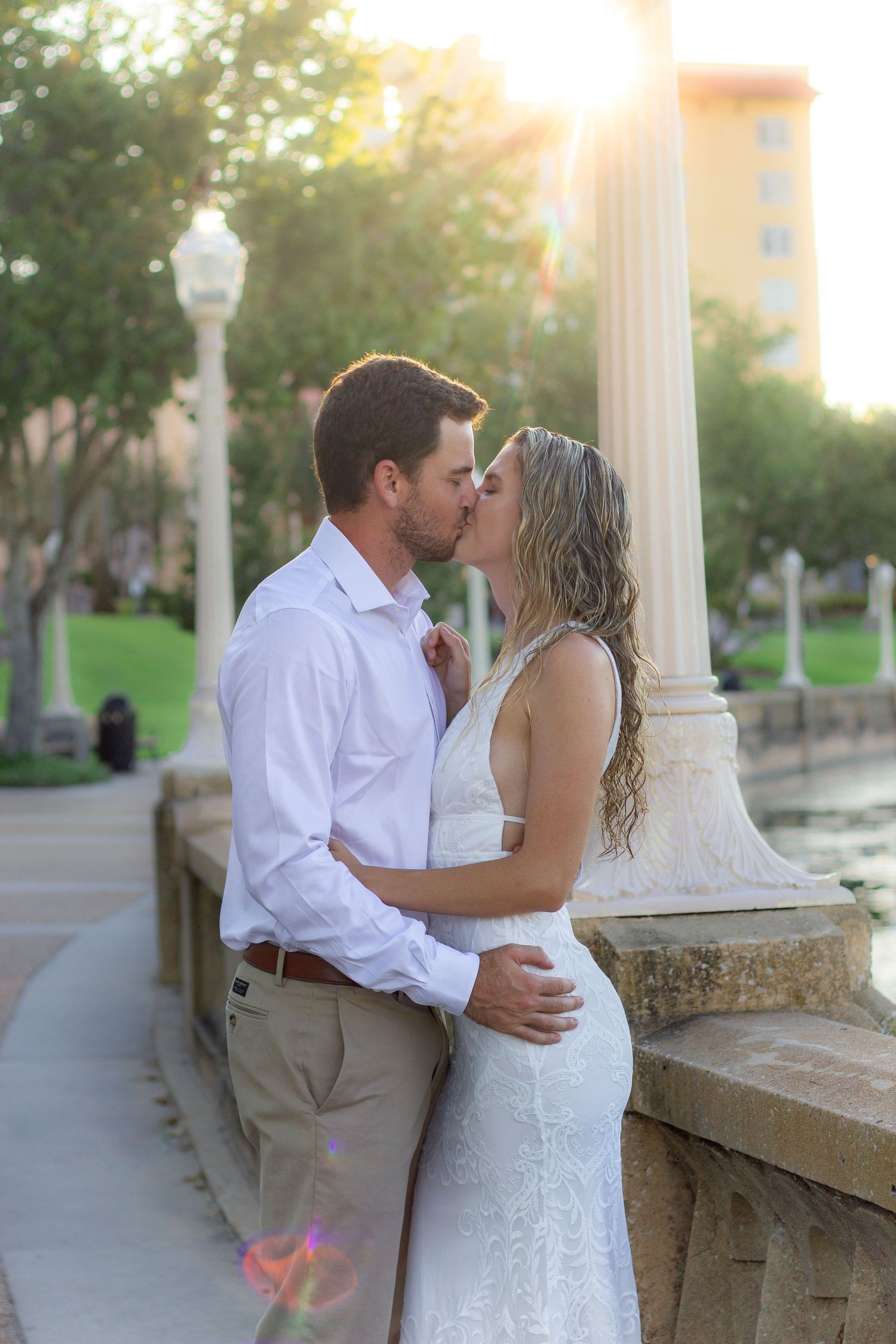 Couple in white dress and dress shirt share romantic kisses at sunset against stone columns and greenery.