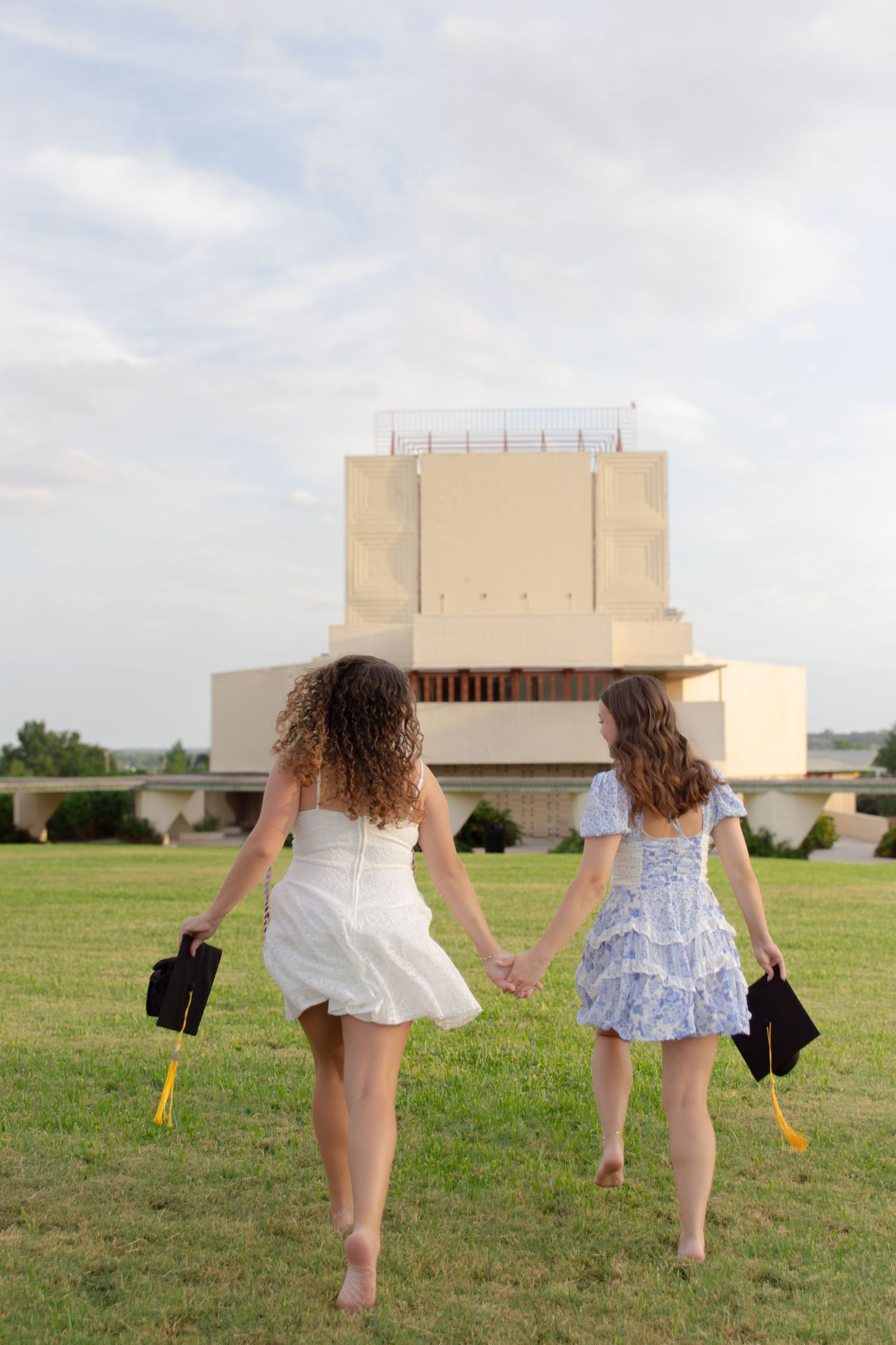 Two graduates in summer dresses walking through a grassy field carrying black graduation caps.