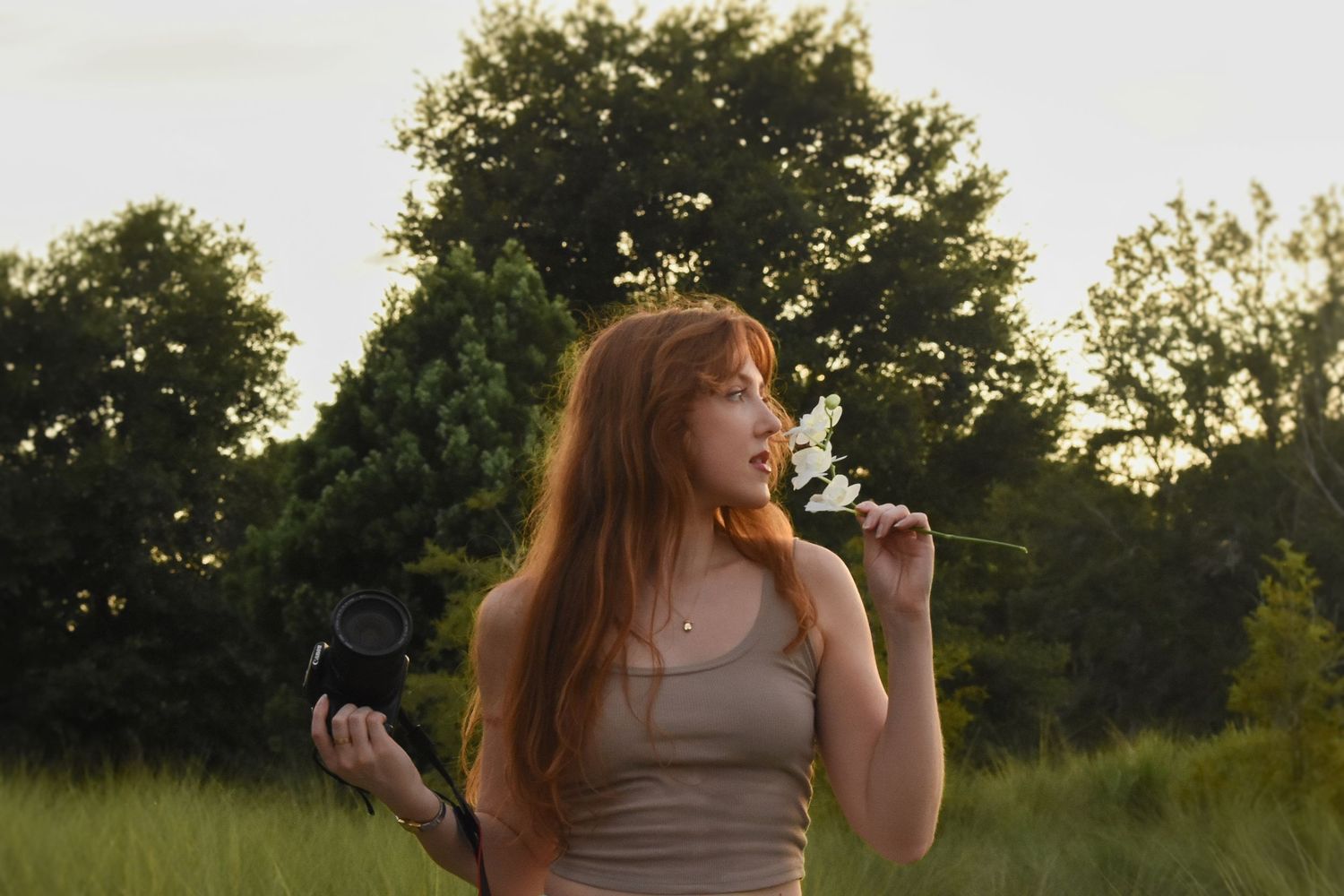 Person in gray tank top poses in a grassy field at sunset with flowers.