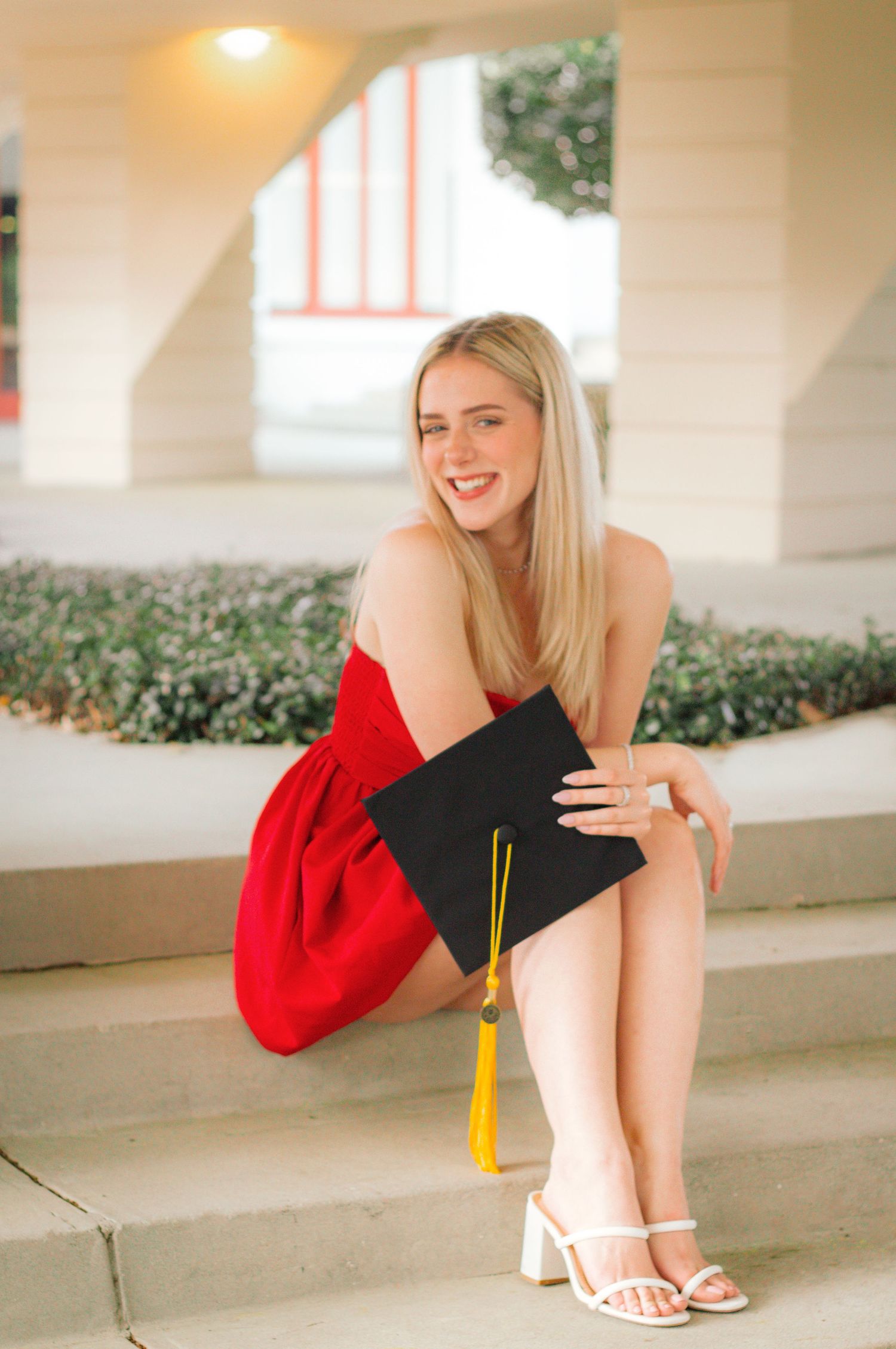 Graduate in red dress sits on concrete steps holding graduation cap with yellow tassel and wearing white sandals.