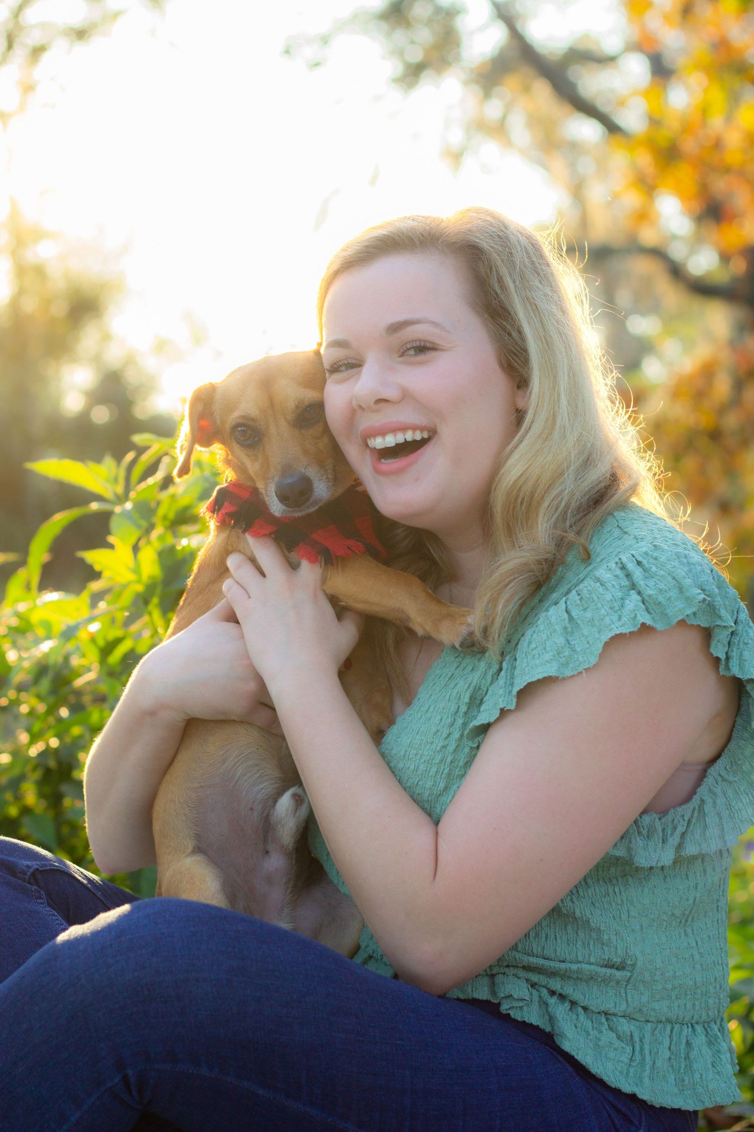 A small brown dog being cuddled outdoors during golden hour with autumn foliage in background.