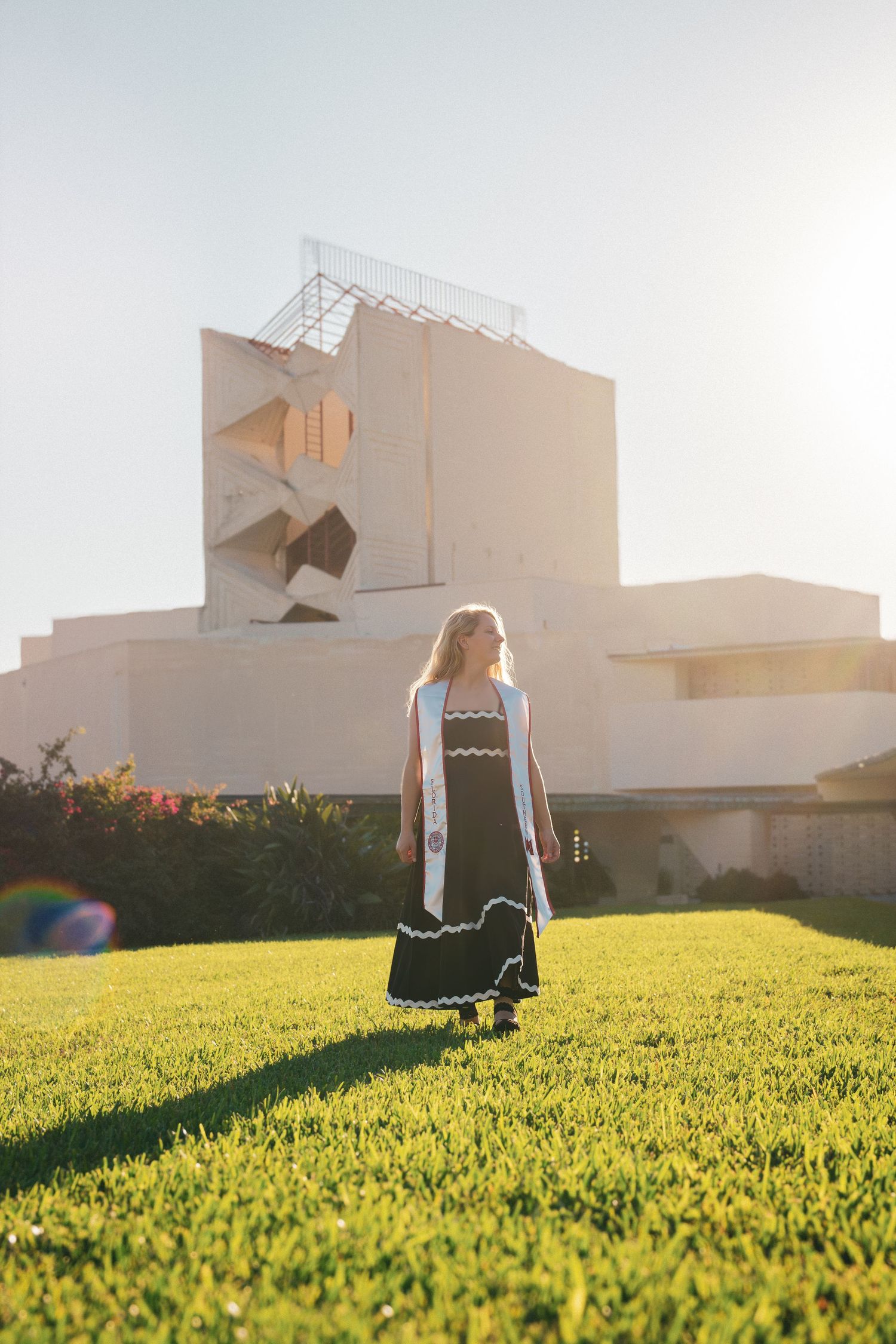 Graduate in black dress walks across sunlit campus lawn at golden hour.