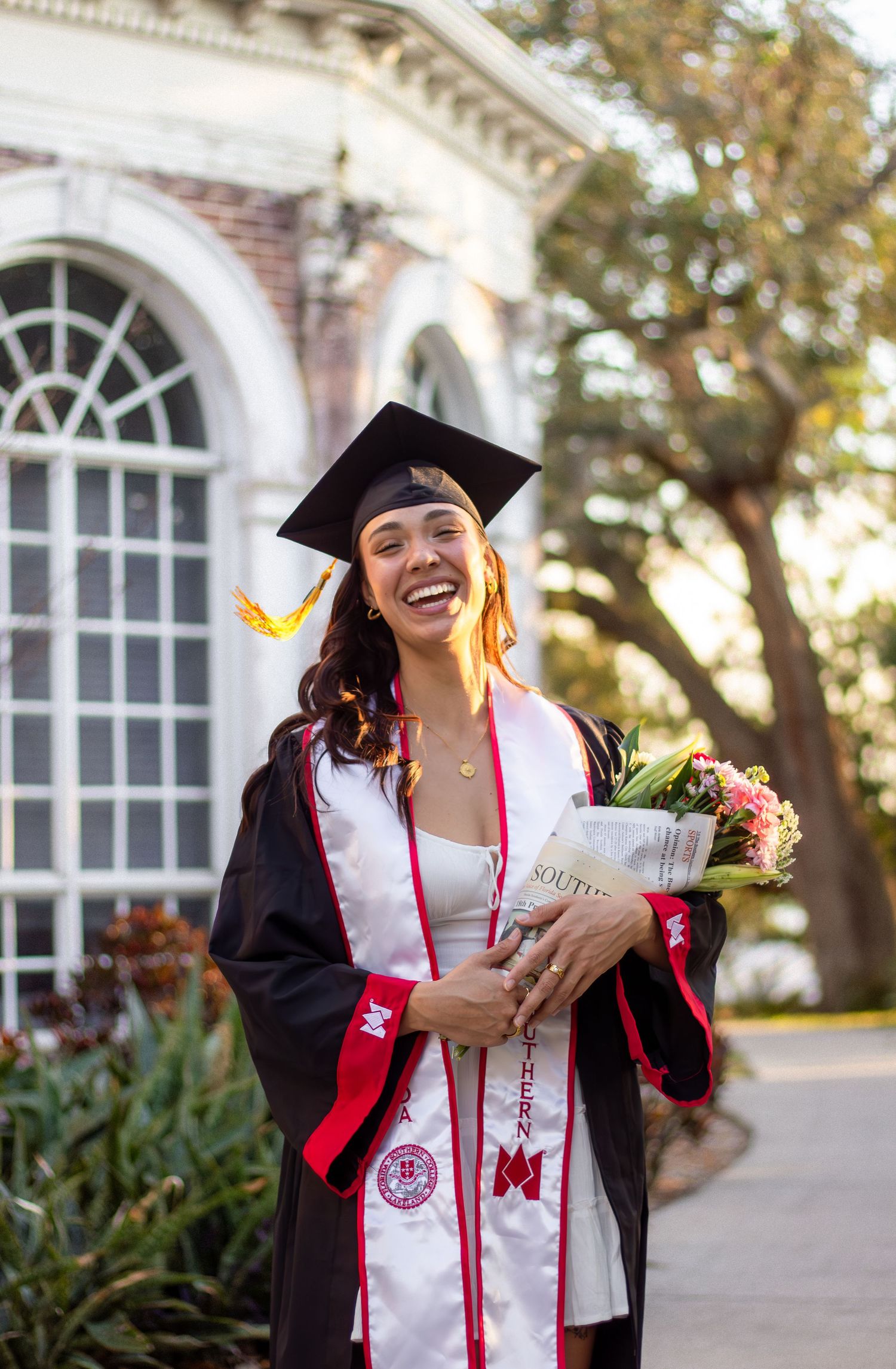 A graduate in cap and gown celebrating on a university campus pathway with architectural arches in the background.