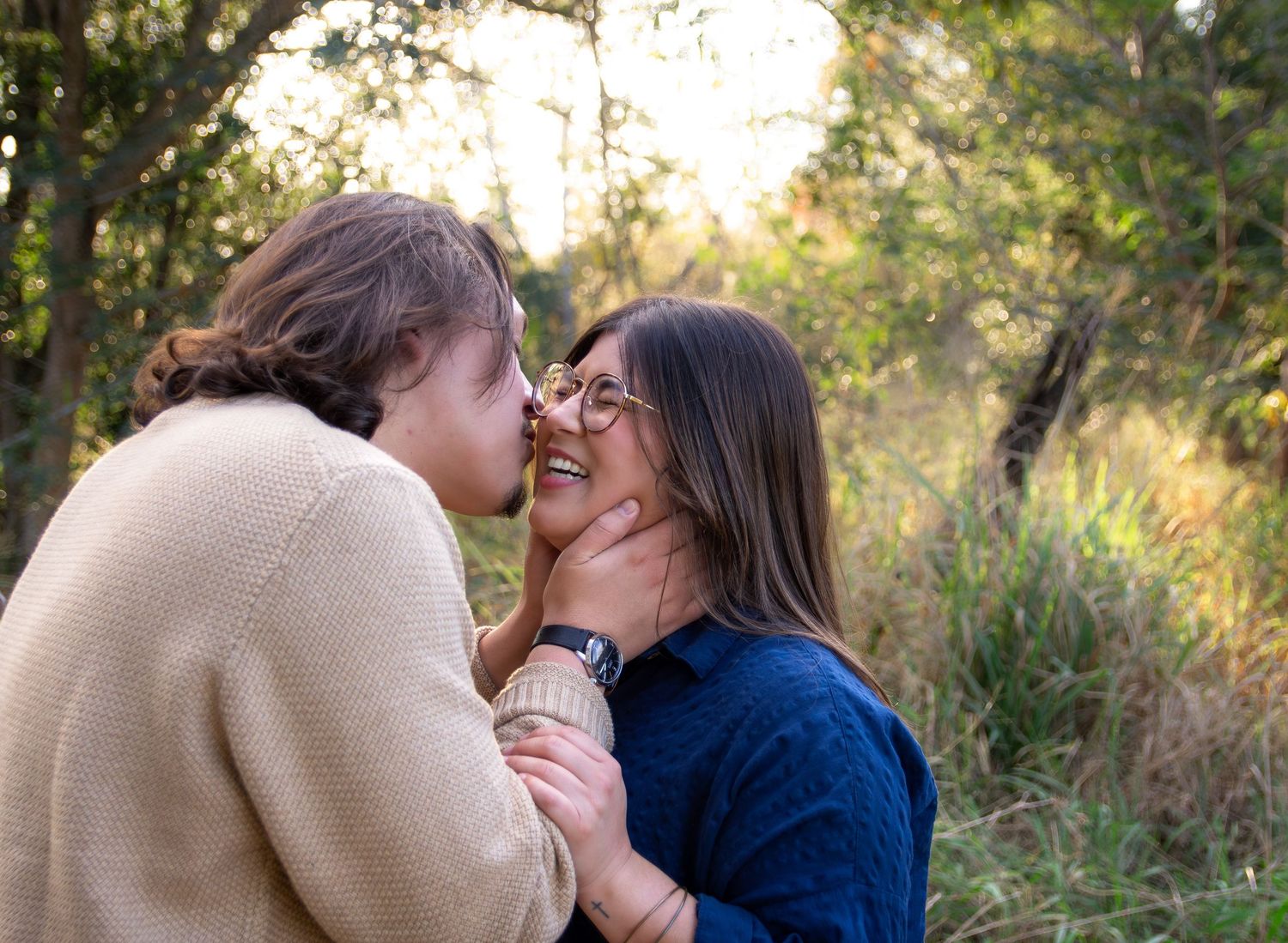 A couple shares a romantic kiss in a natural outdoor setting with golden sunlight filtering through trees.