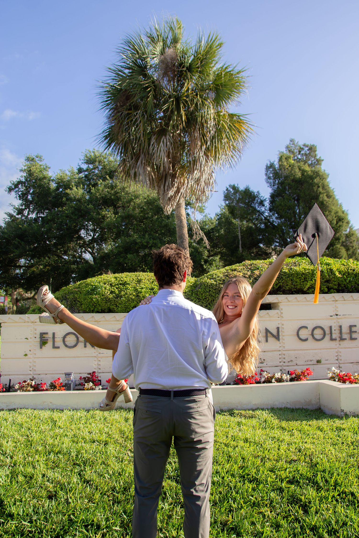 Excited female graduate raises cap and arms in celebration outside Florida Southern College on a sunny day.