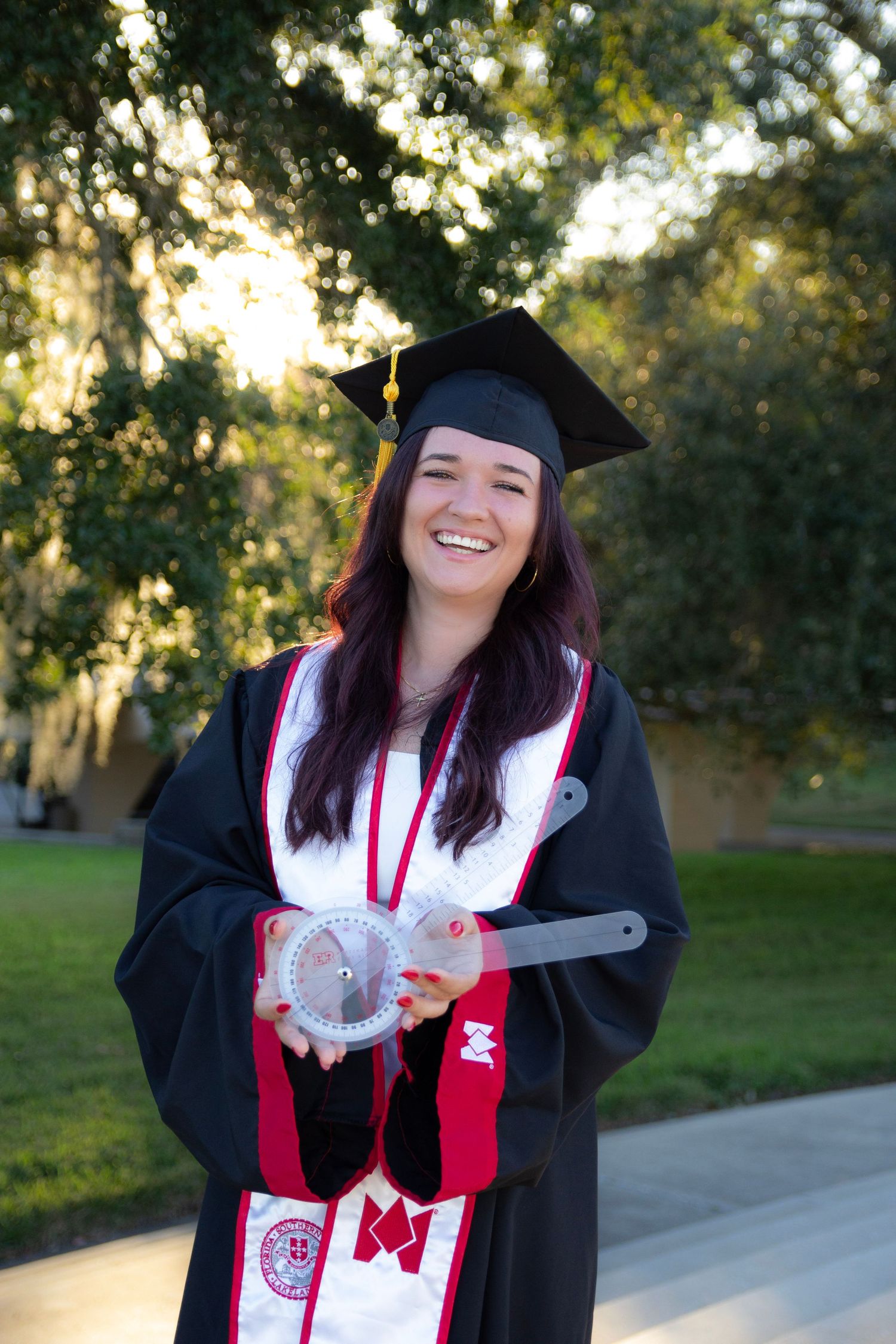 Graduate in black cap and gown with red stole and honor cords poses outdoors during sunset celebration.