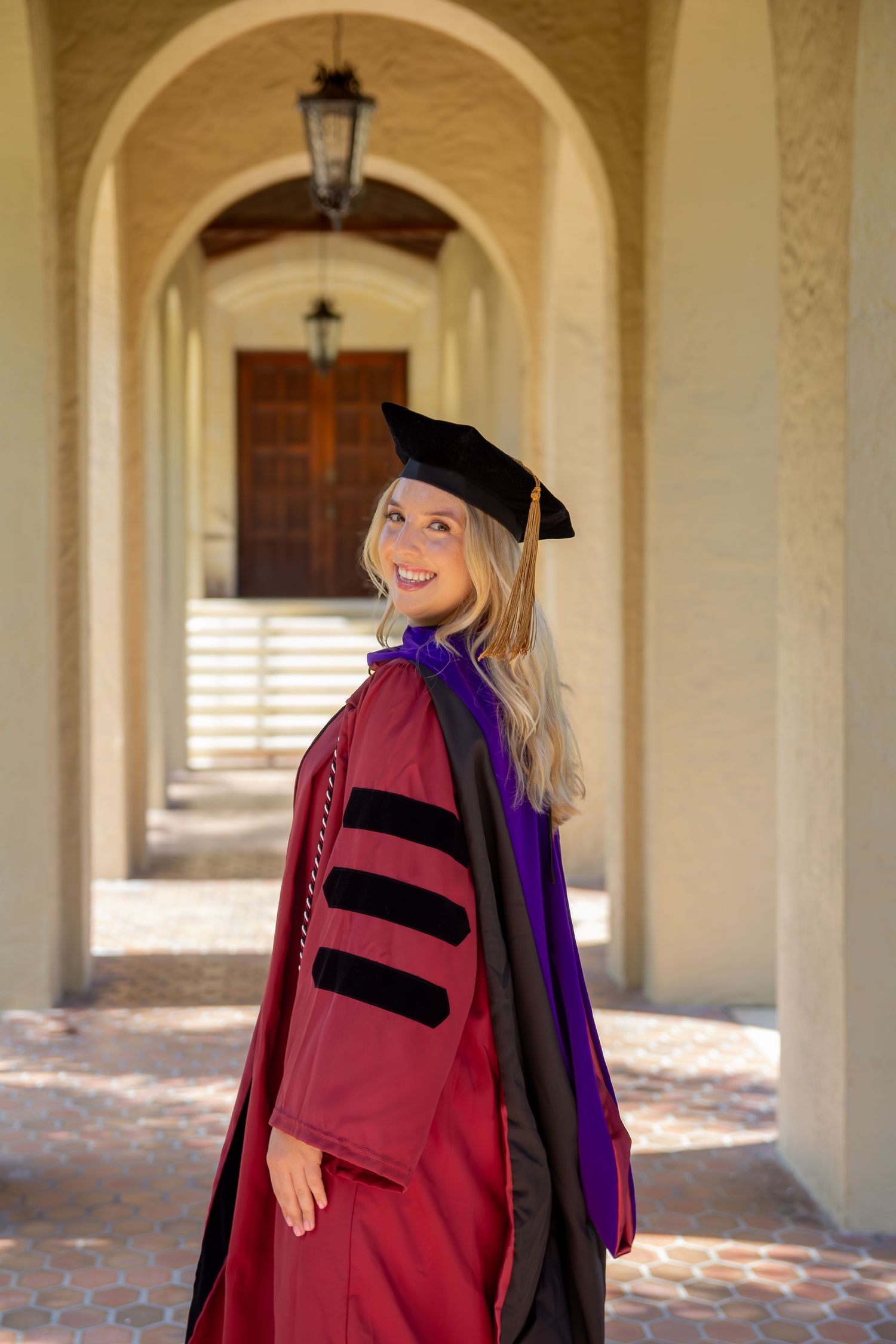 Graduate in red academic regalia posing under ornate archways at university.