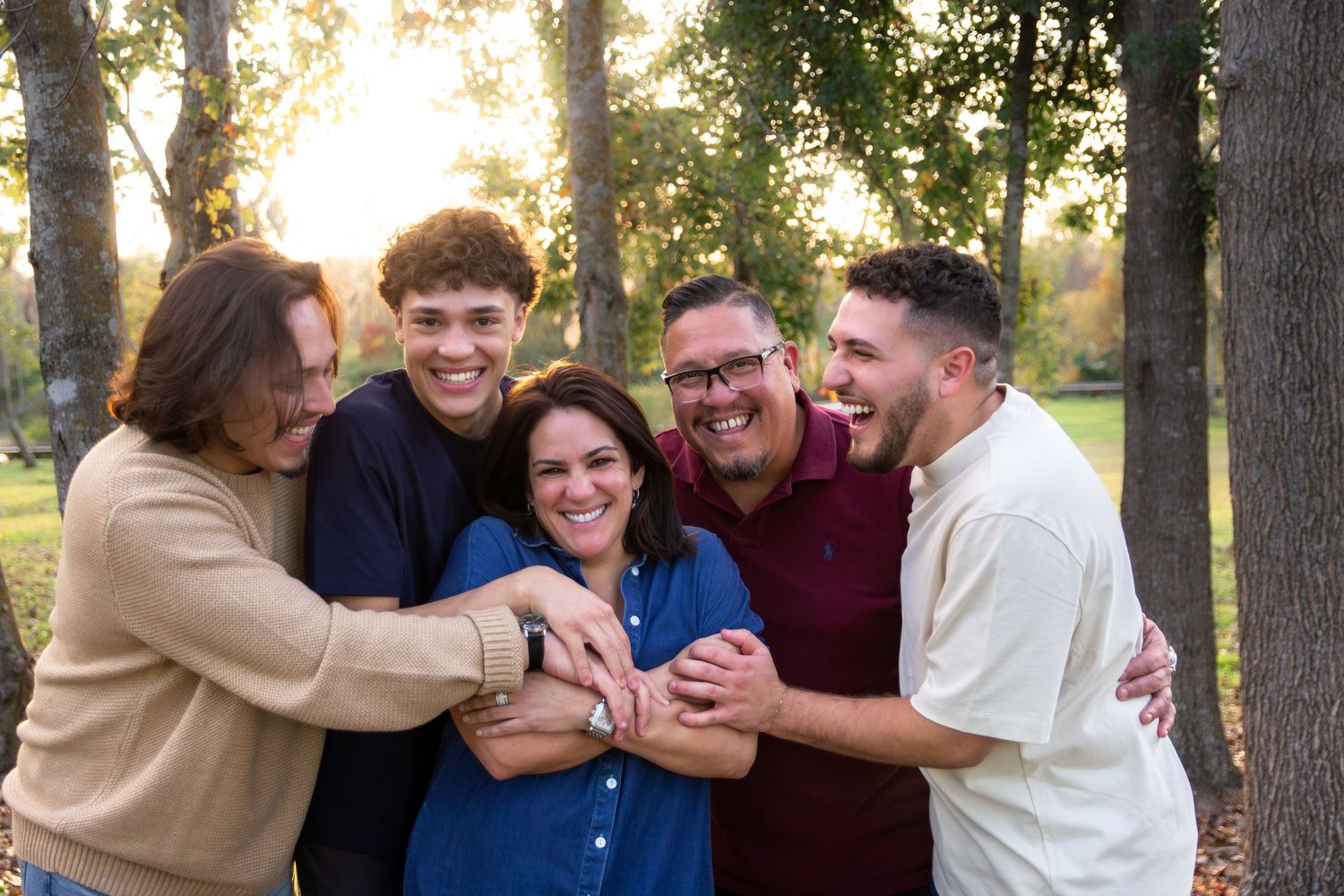 A group of people hugging and laughing together outdoors during golden hour with trees in the background.