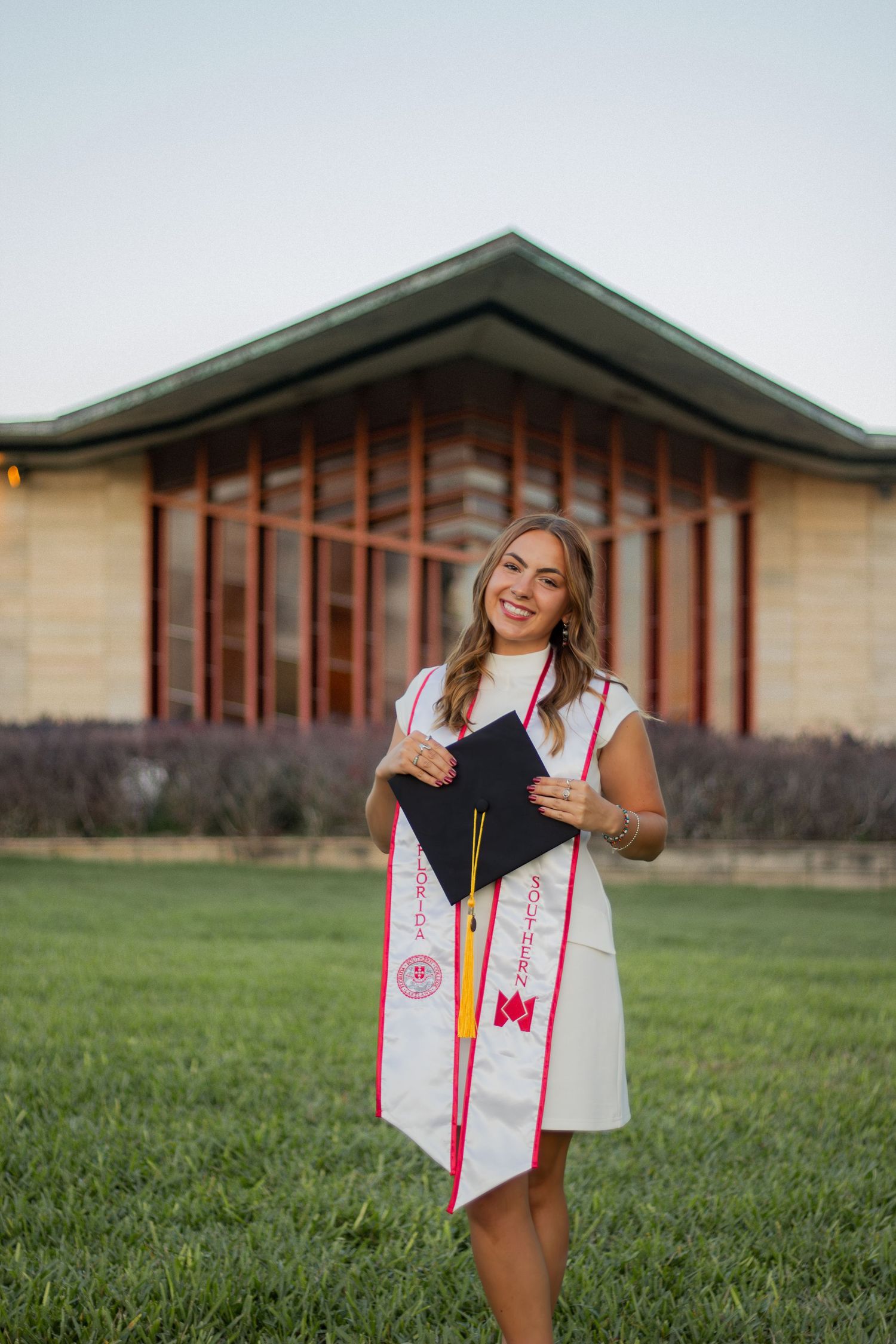 Graduate poses with graduation stoles and decorative sashes on grassy lawn in front of academic building.