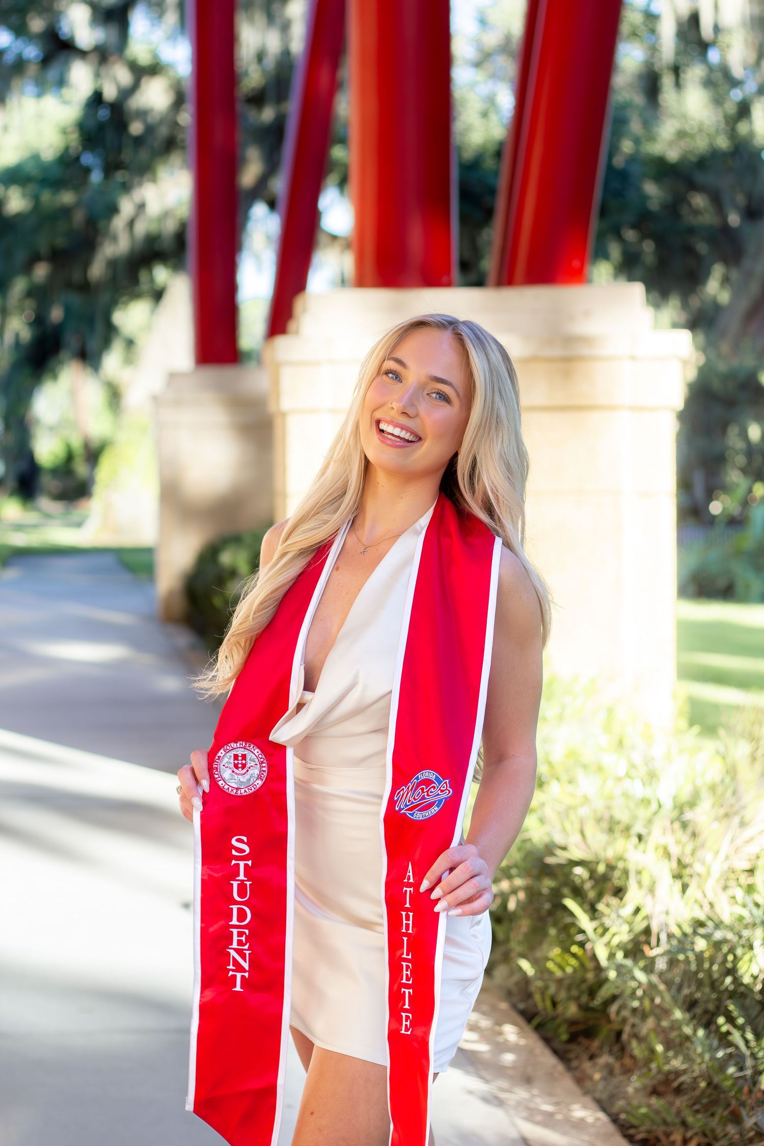Graduate in cream dress poses with red graduation stoles outdoors near red architectural columns.