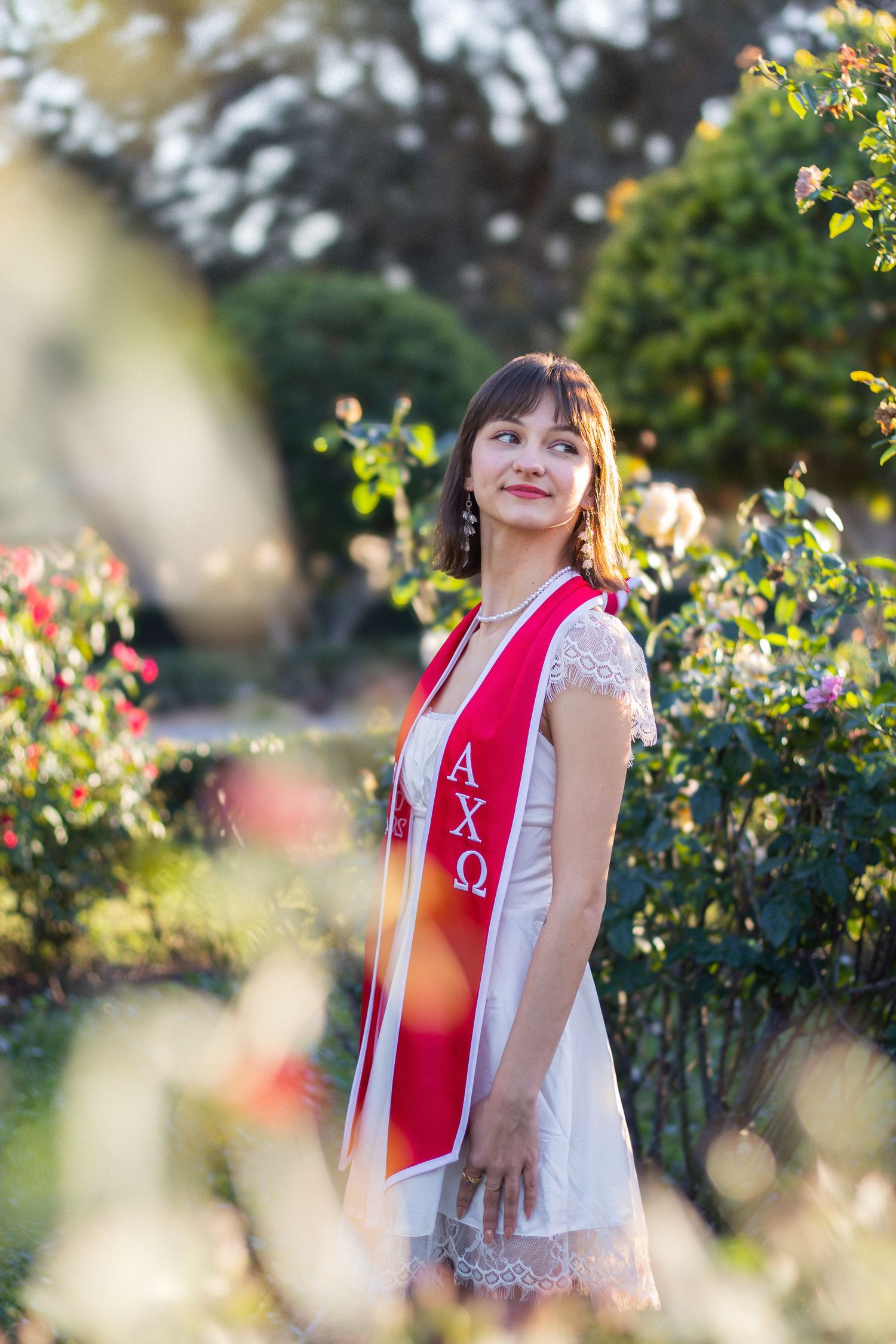 A person wearing a red and white sorority sash poses outdoors in a garden setting with blooming flowers and greenery.