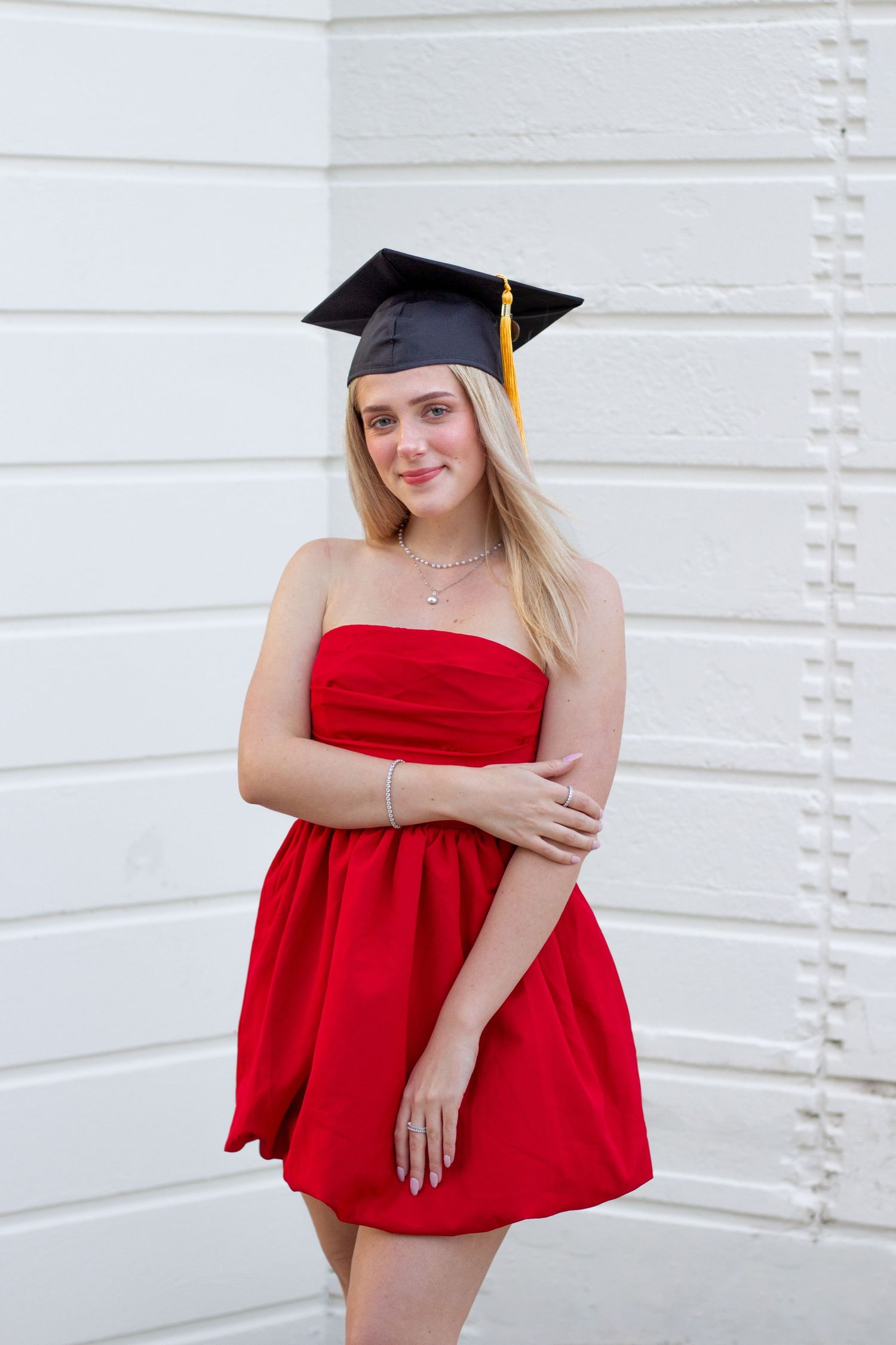 Graduate in red strapless dress and black cap poses against white wall during commencement celebration.