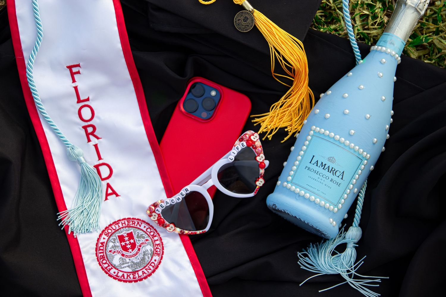 Graduation items including a Florida sash, red iPhone, heart sunglasses, and blue decorative champagne bottle on black fabric.