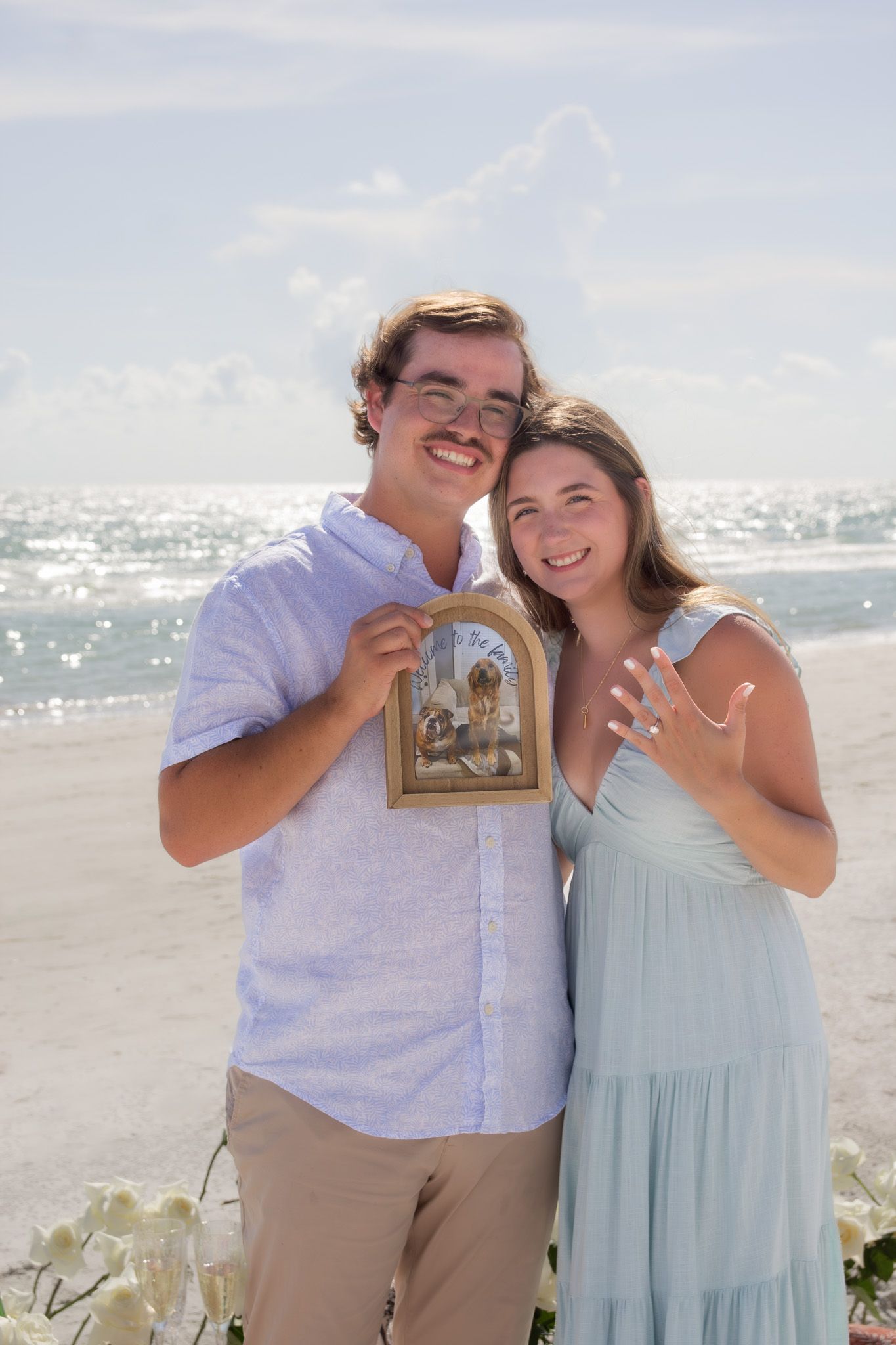 A couple poses playfully on a sunny beach holding up a framed photo.