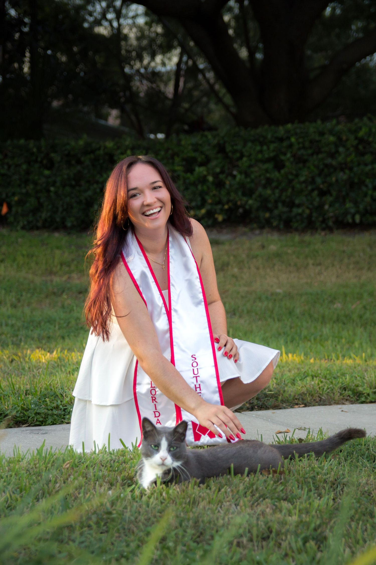 A person in a white dress sits on grass with a gray cat during a sunny outdoor photoshoot.